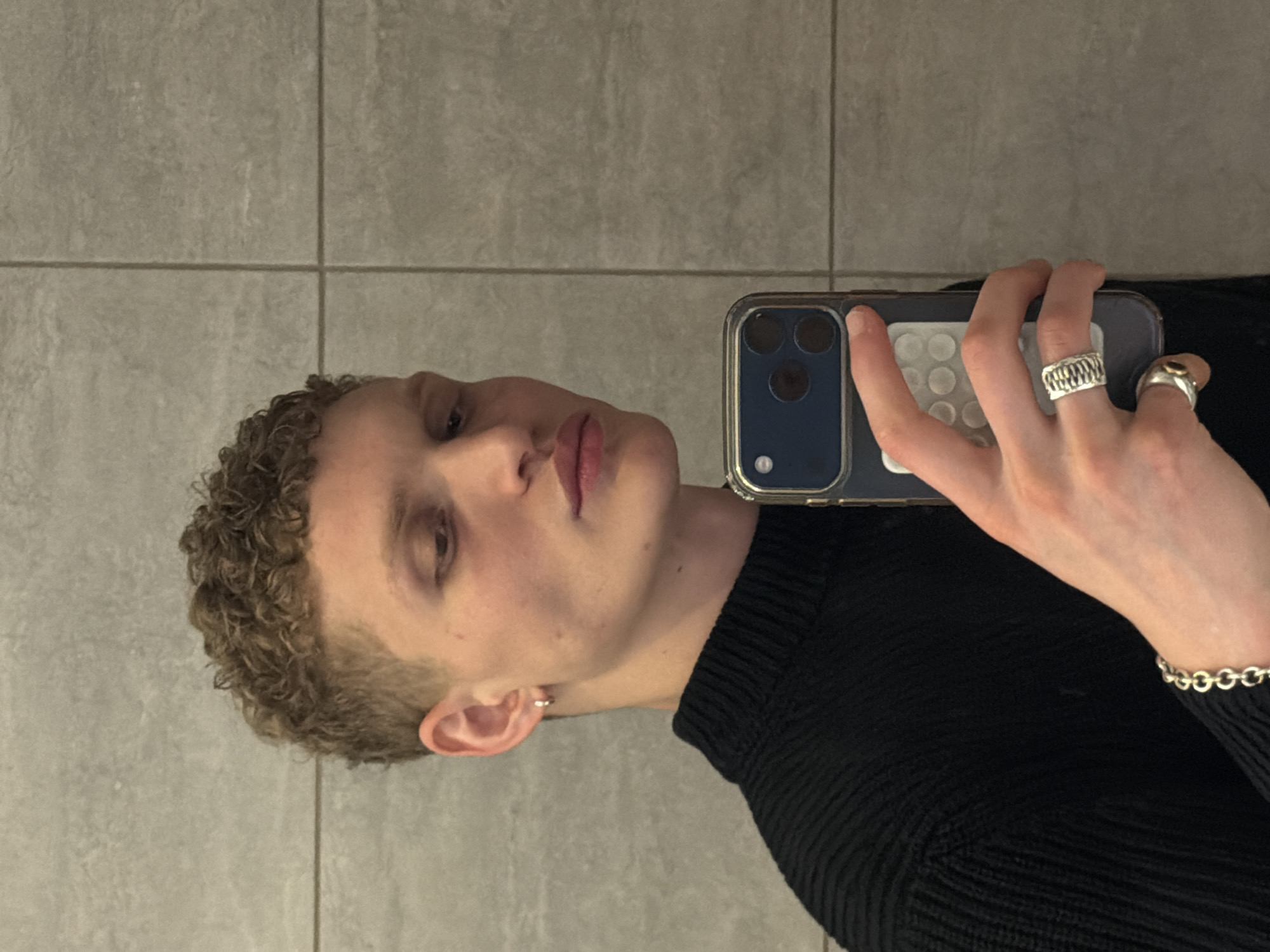 A person with short, curly blonde hair is holding a smartphone in front of their face, presumably taking a selfie. The person is wearing a black sweater and silver jewelry, including rings and a bracelet. The background is a gray tiled wall. No information suggests that the photo was taken in the city of London, United Kingdom.