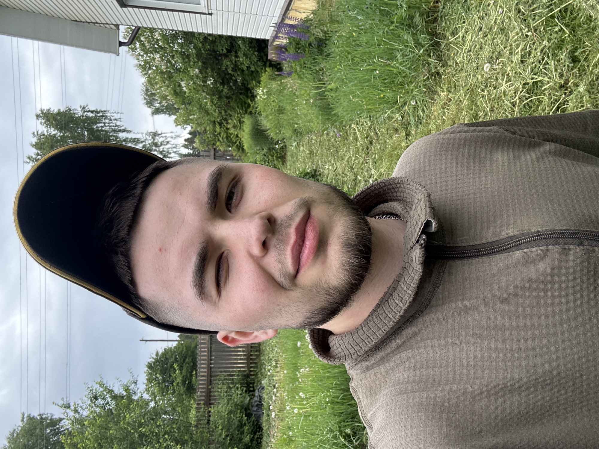 A young man is the central subject of this outdoor portrait, taken in what appears to be a residential or rural setting in Бородулино, Russia. He is positioned prominently in the foreground, looking directly at the camera with a slight smile or smirk, and one eye is partially closed, conveying a relaxed expression. He has a beard and stubble and is wearing a dark cap with a contrasting light-colored edge on the brim. His attire includes a long-sleeved shirt in a khaki or olive-green hue, featuring a textured, possibly waffle-knit, fabric and a zippered collar.
The background reveals an overcast sky, suggesting it is daytime. Elements of the environment include a portion of a house with white siding, a window, and a gutter visible in the upper right. Lush green grass, dotted with wildflowers like dandelions and purple flowers (possibly lupines), covers the ground behind him. Trees and a wooden fence line are also visible, hinting at a backyard or garden area. Faint power lines traverse the sky in the upper left. There is no visible text in the image.