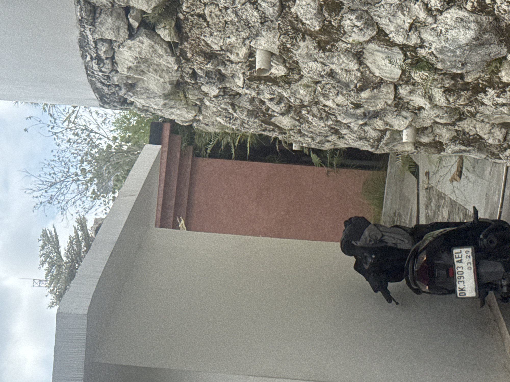 The image shows a paved walkway between two structures. On the left is a light-colored building with a curved edge, and on the right is a wall constructed of rough, grey stones. A dark-colored scooter is parked on the walkway, facing away from the viewer. Clothing is draped over the handlebars and seat of the scooter. A tall, reddish-brown rectangular structure is visible between the two buildings. Vegetation, including ferns, grows in the gap between the reddish structure and the stone wall. The sky above is overcast. The license plate on the scooter reads "DK.3903 AEL 03-29". The scene is likely in the city of Kuta Selatan, Indonesia.