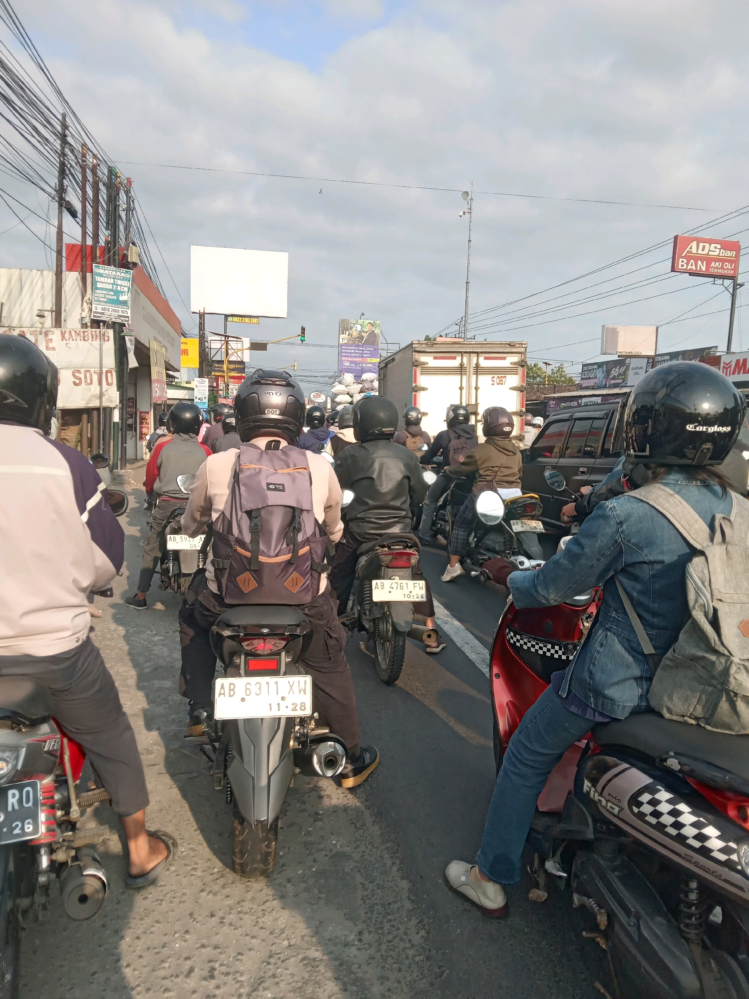 The image depicts a street scene in Sitimulyo, Indonesia, characterized by heavy motorcycle traffic. In the foreground, the backs of several motorcyclists are visible, most wearing helmets and backpacks. One individual, in the center right, wears a denim jacket and a grey backpack, riding a red scooter. To their left, another person in a grey jacket and backpack is on a motorcycle with a license plate reading "AB 5917 AF". Further left, another motorcyclist in a light-colored jacket is seen from behind. In the background, a white billboard stands prominently, alongside various signs and utility poles with numerous overhead wires. Commercial buildings line the street, with more motorcycles and vehicles visible further down the road, suggesting a busy urban environment. The sky is partly cloudy, indicating daylight.