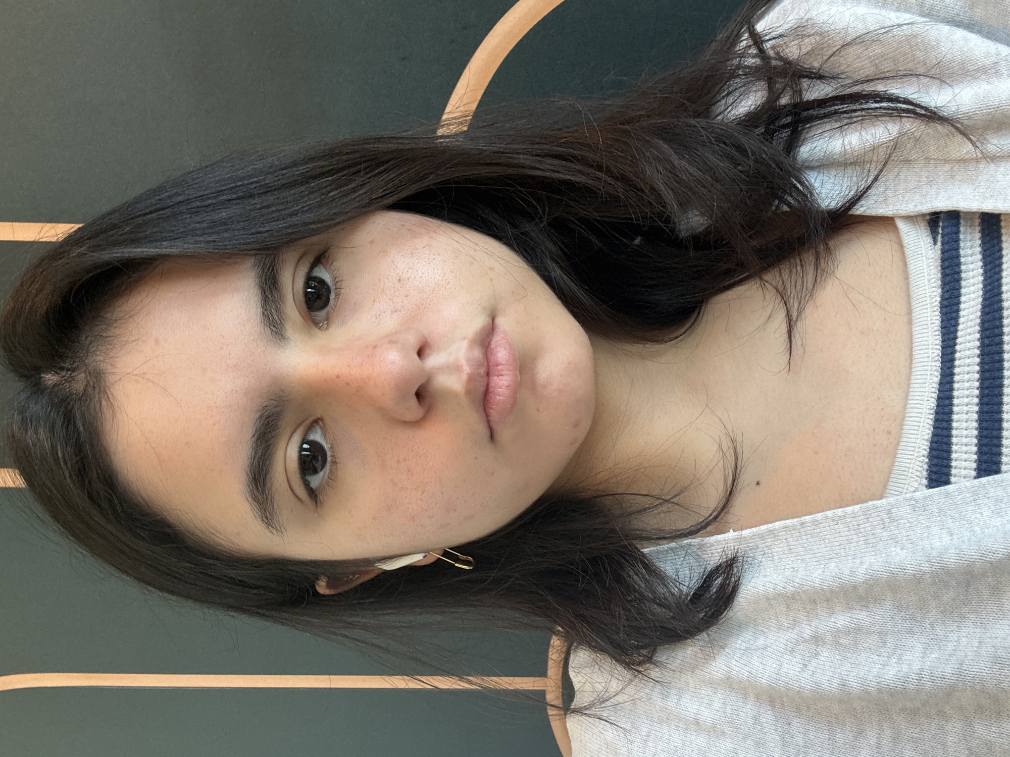 A young woman is centered in the image. She has dark brown hair and eyes, and freckles on her face. A gold safety pin is affixed to her left ear. She wears a light gray top with visible blue and white stripes on the neckline. The background behind her is a dark green wall with light brown wood trim. The photograph was taken indoors, but the specific location in Austin, United States of America is not identifiable from the image.