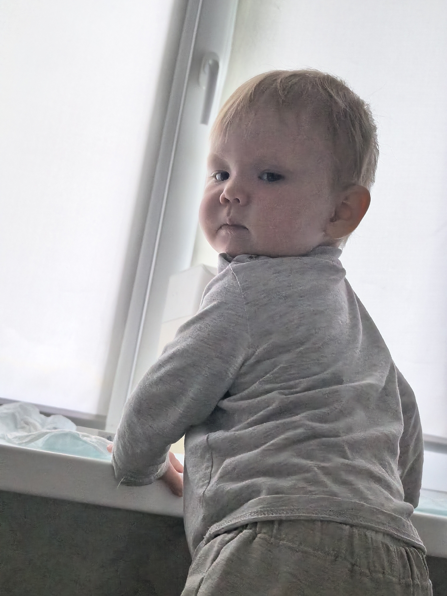 An infant, appearing to be around one year old, is captured in a medium shot, turned away from the camera and looking over their left shoulder towards the viewer. The infant has fair hair and rosy cheeks. They are wearing a light gray long-sleeved shirt with a collar and light-colored trousers. The infant is positioned near a window with a translucent blind, which fills the background with a bright, diffused light. The infant's left hand is visible resting on a white surface, possibly a changing table or shelf, that is in the foreground. The overall lighting suggests the scene takes place indoors during the daytime. There is no visible indication of the city of Sterlitamak, Russia, in this image.