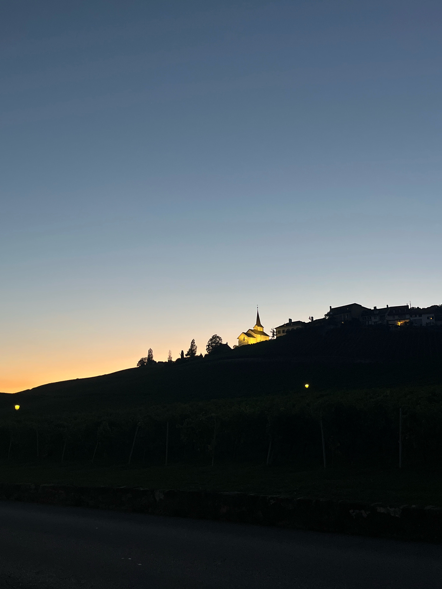 The image depicts a twilight scene over Féchy, Switzerland, featuring a village perched on a hillside. The sky transitions from a deep blue at the top to a soft orange glow near the horizon, indicative of sunset or sunrise. A prominent church with a steeple, illuminated by warm yellow light, stands out on the crest of the hill. Adjacent to the church are several houses, also showing interior lights. The silhouette of trees and the dark shape of the hill are visible against the lighter sky. Below the village, rows of vineyard trellises are faintly outlined in the foreground, with a few streetlights casting yellow glows. A paved road or path runs along the bottom of the frame, with a low stone wall bordering the vineyard. No people are visible in the scene.