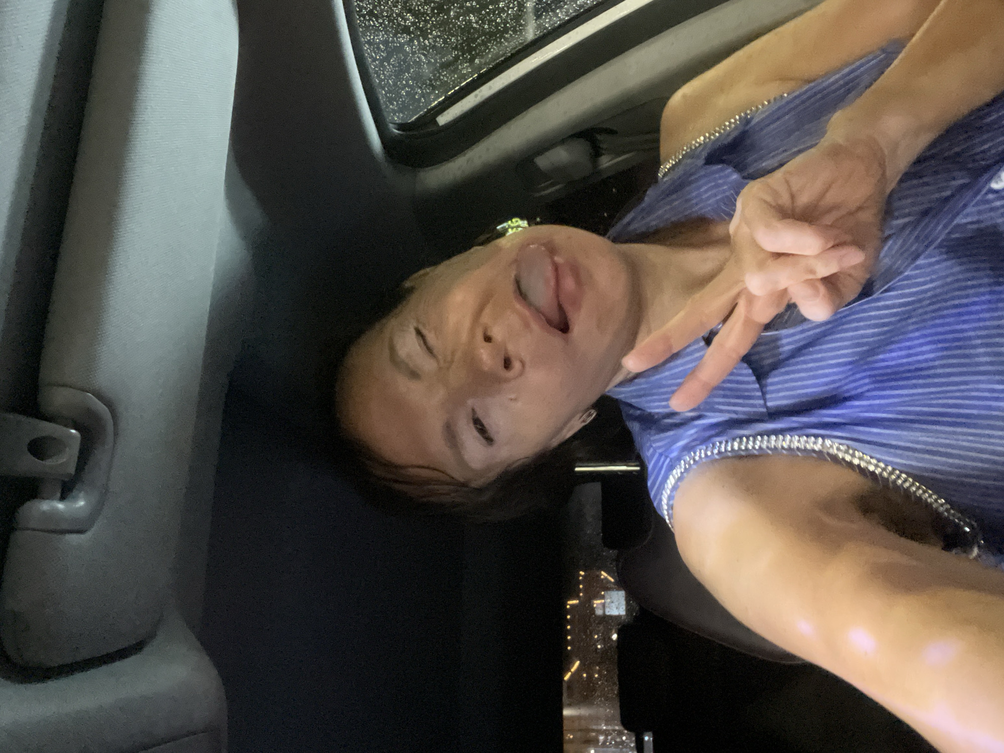 A woman is in the driver's seat of a car at night. She is looking at the camera and making a peace sign with her right hand. It appears to be raining outside as there are water droplets on the car window. The woman is wearing a blue and white striped shirt with silver trim on the straps.