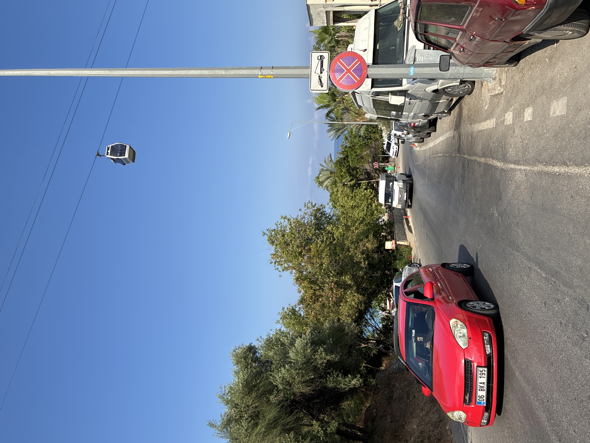 A sunny, clear day is depicted in Alanya, Turkey, featuring a road with vehicles and a cable car. In the foreground, a red car with license plate "06 BKA 195" is driving on the right side of the road. To the right, several parked vehicles, including a white van and a red sedan, line the side of the road. Traffic signs are visible on a pole, one indicating towing and another a red "X" symbol, signifying no parking or stopping. Trees and lush vegetation are on the left side of the road, with the sea visible in the background. A cable car is suspended in the sky, moving across the frame. The overall scene is one of a typical Mediterranean coastal town.
