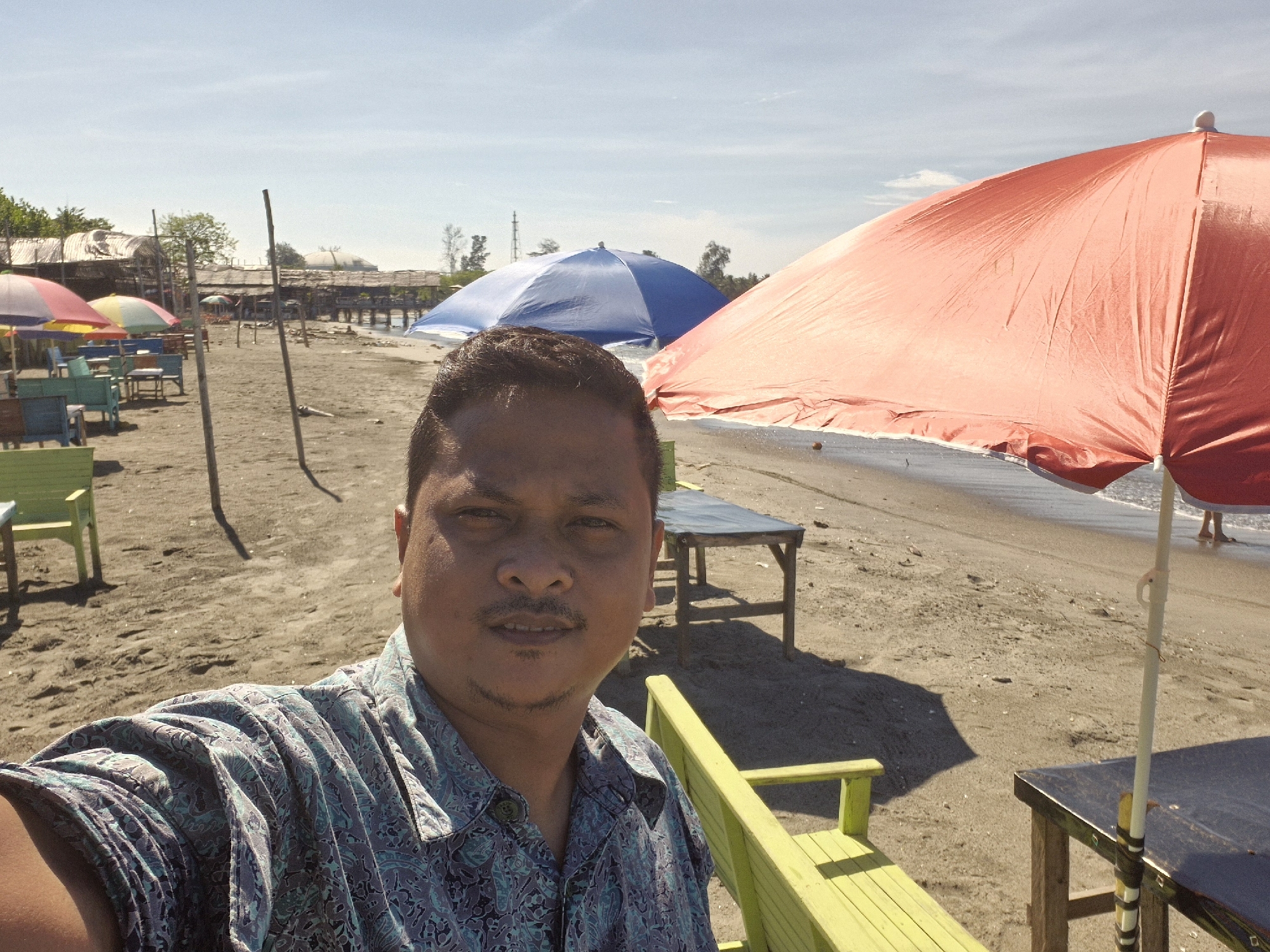 The image shows a man taking a selfie at a beach. He is wearing a patterned blue shirt. Behind him, the beach is populated with tables and chairs, most of them painted in various colors. Several beach umbrellas in red and blue offer shade. In the distance, there are structures built on stilts over the water. The location is Lhokseumawe, Indonesia, suggested by the beachside structures and vegetation. The overall atmosphere is sunny and relaxed.