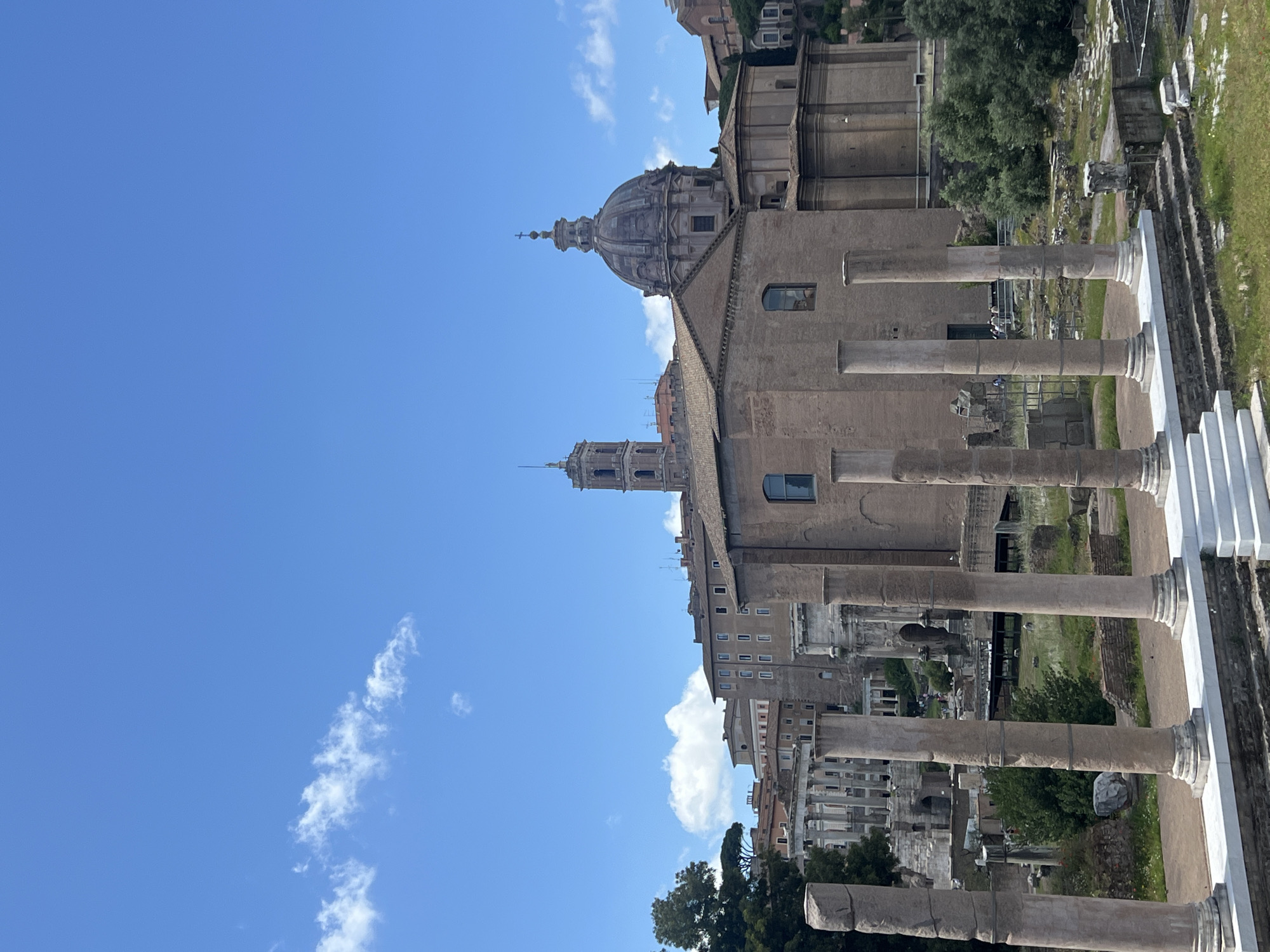 The image captures a view of the Roman Forum in Rome, Italy. The foreground shows partially preserved columns and ancient ruins. In the background, there's a large building with a dome and a tall tower structure. The sky is blue with scattered clouds. The overall scene is of a historical site.