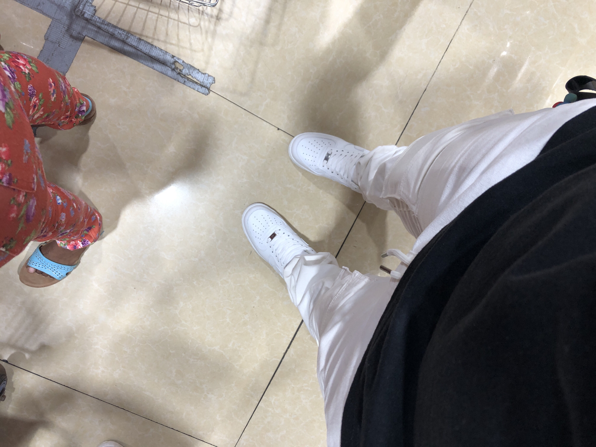 The image shows a view of two people's lower bodies and feet in what appears to be an indoor location with tan-colored tile flooring. To the left, a person is wearing a floral print pajama pant and sandals. To the right, another person is wearing white sneakers, white pants, and a black garment. A shopping cart is visible partially in the upper left of the frame. The location in the city of Juan Griego, Venezuela cannot be ascertained from this image alone.