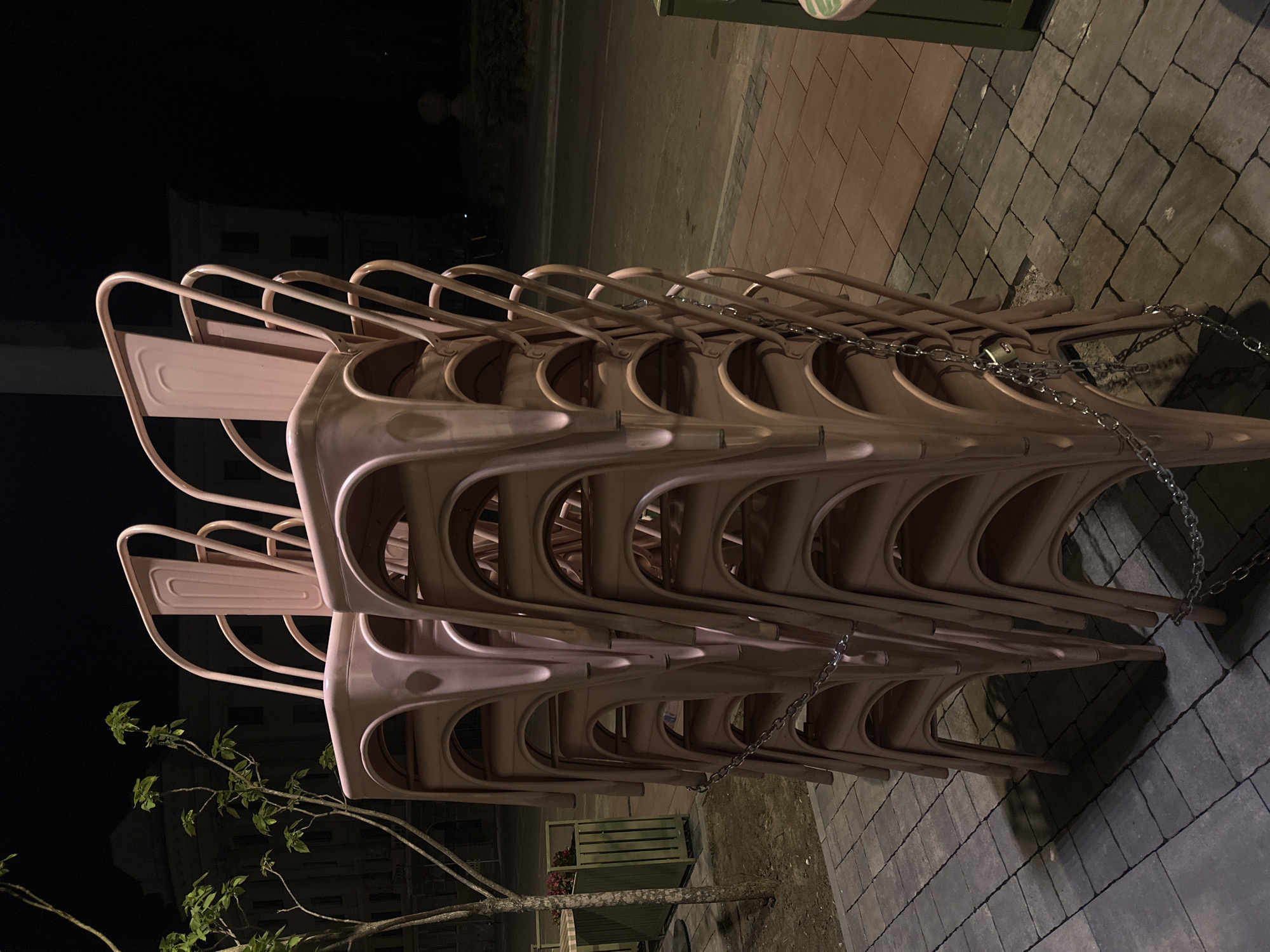 A stack of light pink metal chairs, secured by a silver chain, sits on a paved surface. The pavers are predominantly gray, with some sections of reddish-brown brick. In the background, on the left, a tree branch with green leaves is visible, along with a dark building. The scene appears to be lit artificially, and the background is dark, suggesting nighttime. It is plausible this image was taken in Ternopil, Ukraine.