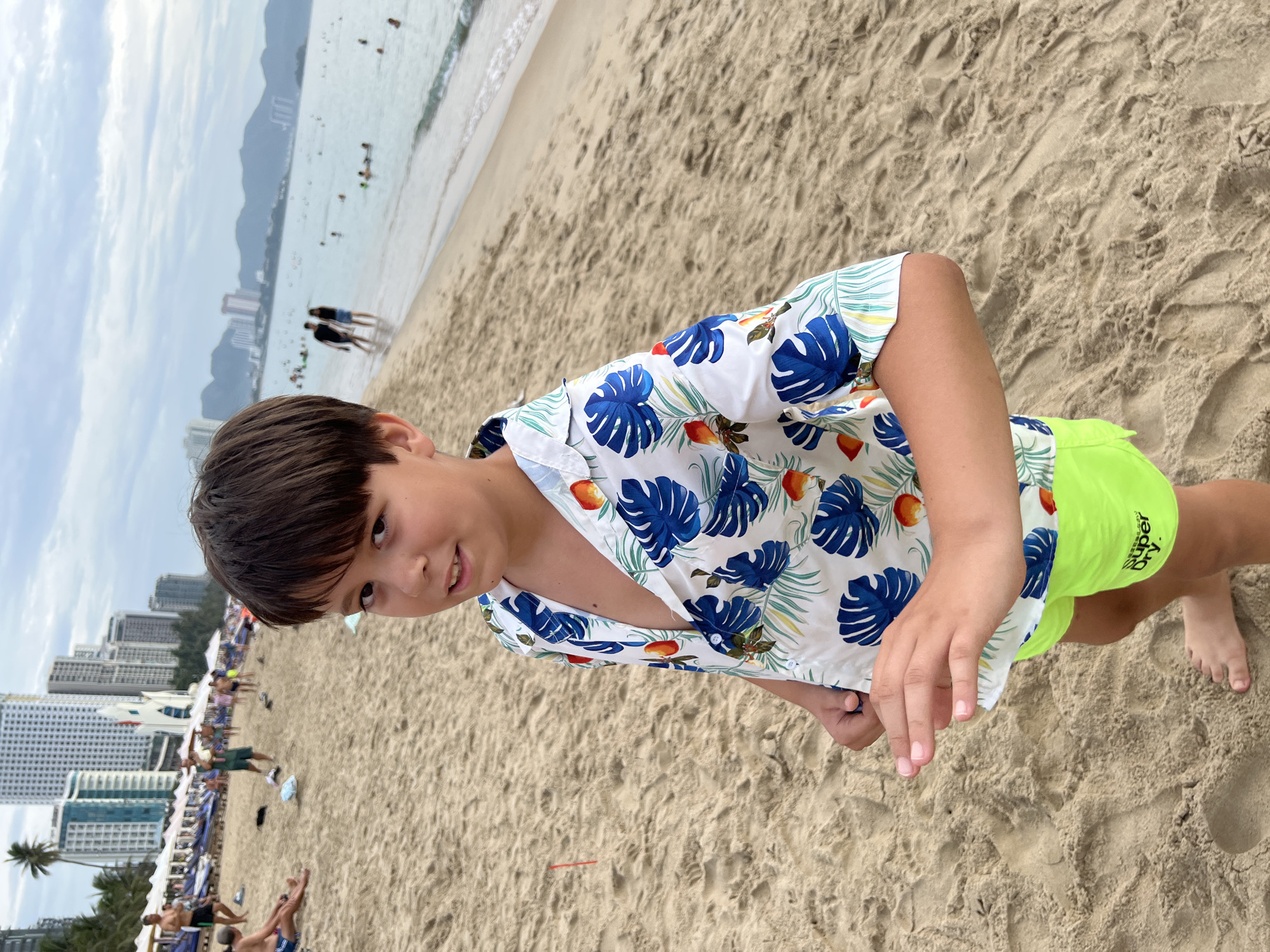A young boy stands on a sandy beach in Nha Trang, Vietnam, looking at the camera. He is wearing a white short-sleeved shirt with a tropical pattern of blue leaves and colorful fruit, and bright green swim shorts. His right hand is held up, fingers spread. In the background, the ocean stretches to the horizon with distant hills and buildings. Other beachgoers are visible in the distance, some in the water and some relaxing on the sand. The sky is overcast.