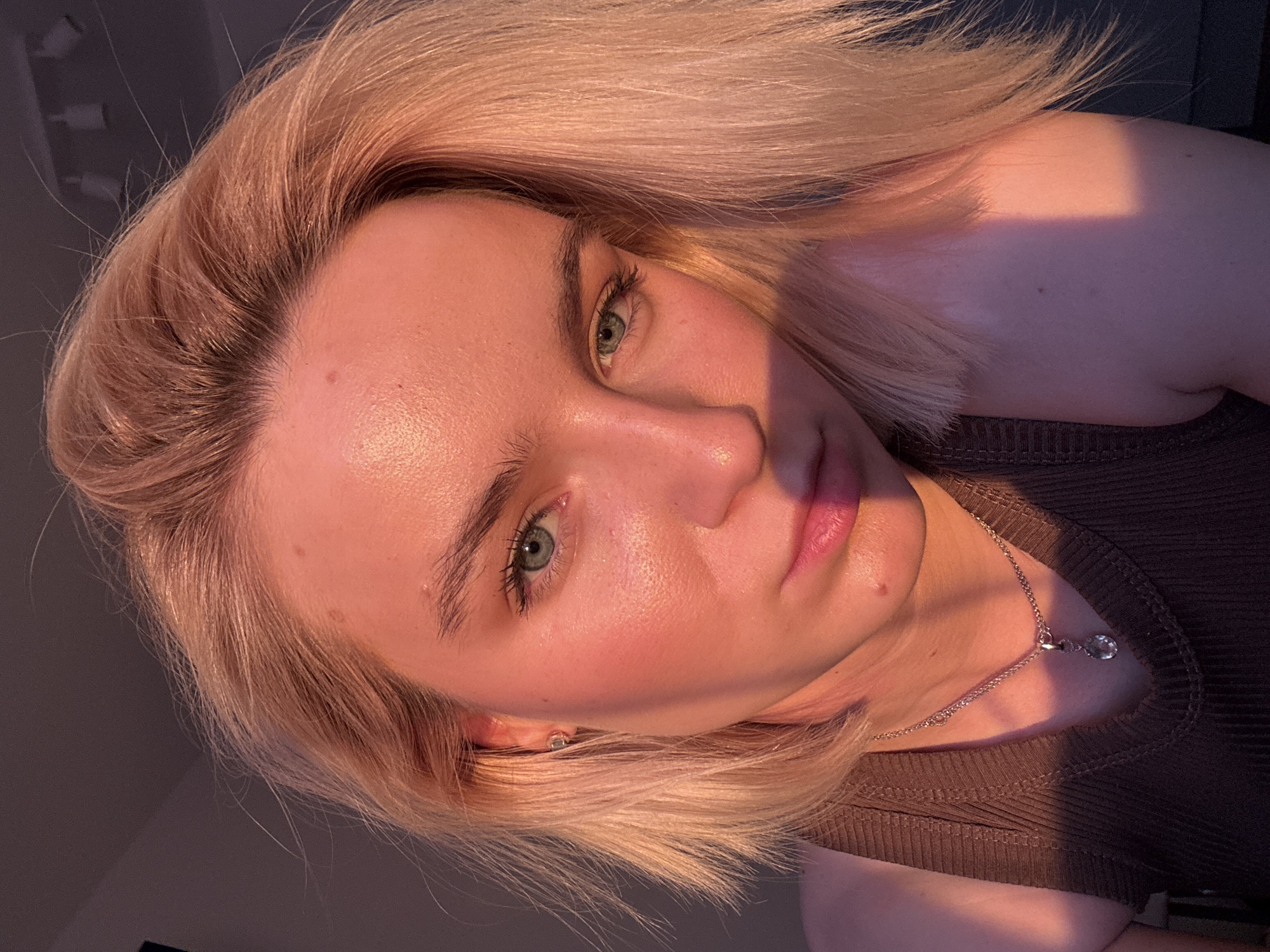 A close-up selfie shows a woman with blonde, shoulder-length hair styled in a blunt cut. She has fair skin and green eyes, and her face is partially illuminated by warm, golden light, casting shadows on her cheek and neck. The light suggests a late afternoon or early evening setting. She is wearing a dark, ribbed tank top and a delicate silver necklace with a pendant. The background is dark and out of focus. The image provides no evidence of a location in Vsevolozhsk, Russia.