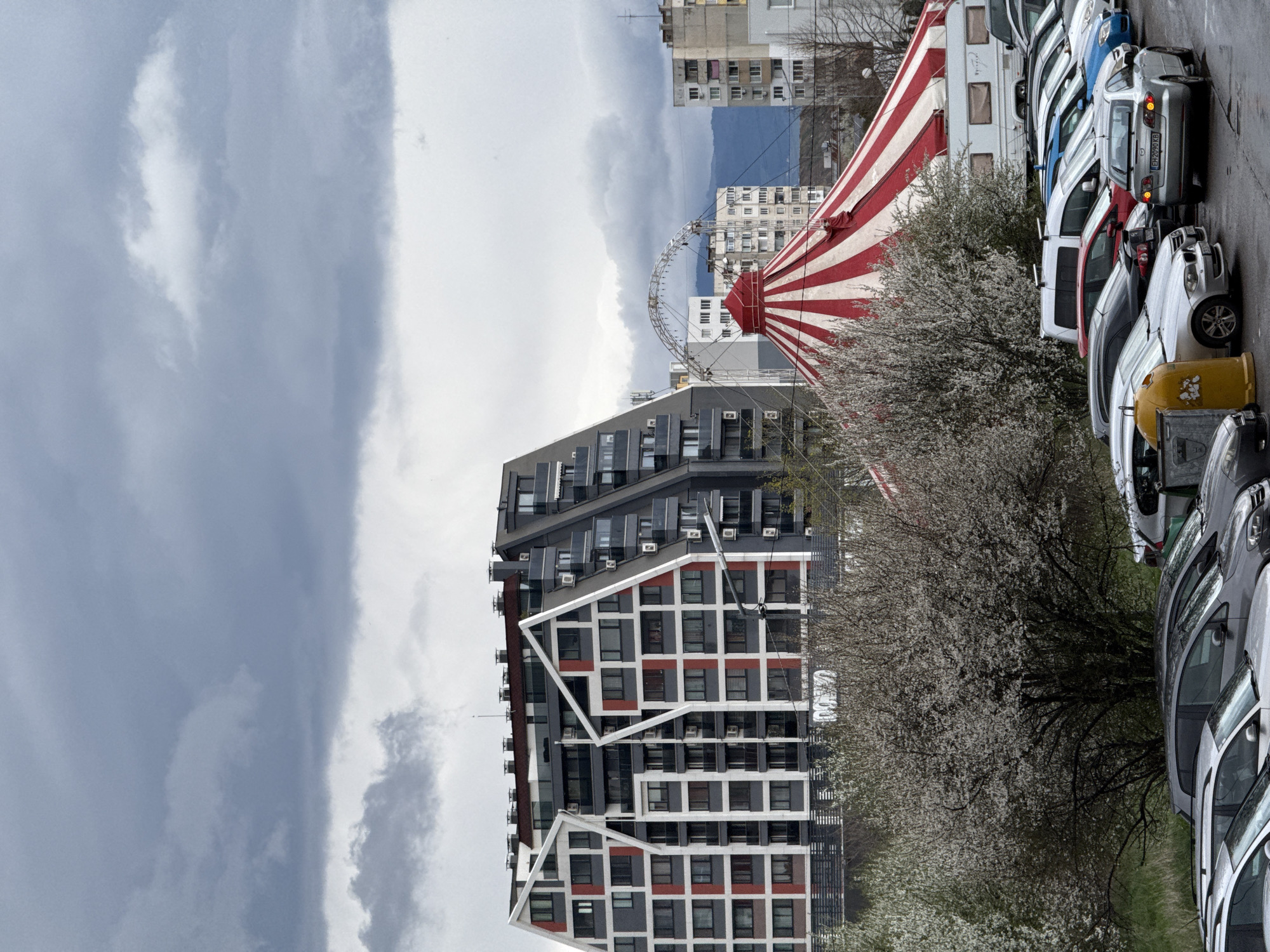 The image depicts an urban scene in Sofia, Bulgaria. A modern, geometrically designed building with alternating gray, white, and red patterned facades stands prominently in the frame. Adjacent to it is a circus tent, marked with red and white stripes, next to a Ferris wheel. Blooming trees are scattered around the scene, adding a touch of nature. A row of parked cars lines the street, further indicating the urban setting. In the background, apartment buildings and a cloudy sky can be observed. The overall impression is a mix of modern architecture, recreational elements, and typical urban infrastructure.