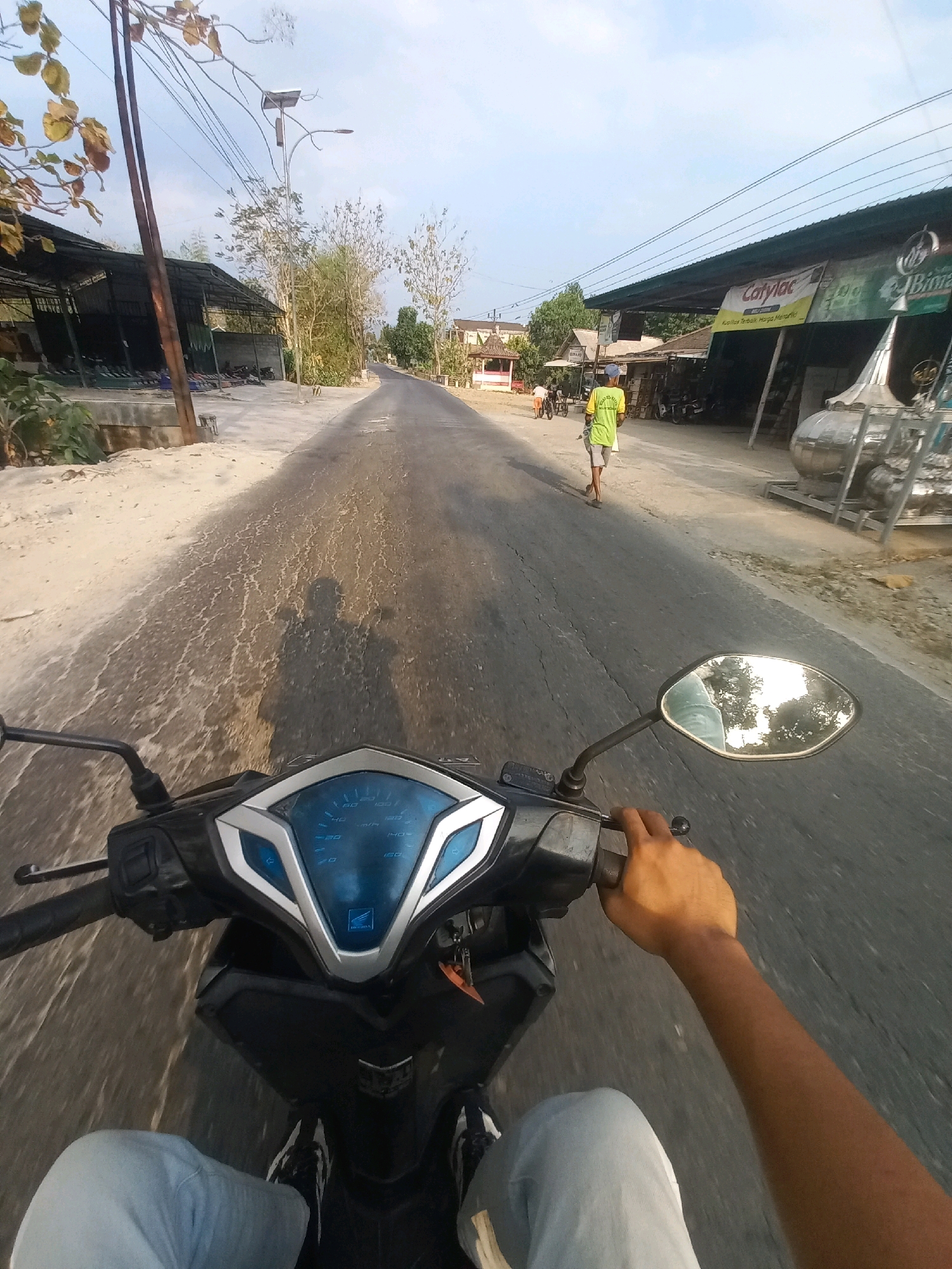 The image captures a first-person perspective from a motorcycle traveling on a paved road in Ngarjosari, Indonesia. The road surface is cracked and weathered, with patches of lighter colored material on the left side. To the left of the road, a long, open-sided structure with a corrugated metal roof is visible, beneath which are rows of what appear to be mats or sleeping areas. A solar-powered street lamp stands near the road. Further down the road, on the right side, a row of shops or businesses are situated, one of which has a prominent "Catylac" sign. Several people are visible in the distance: a person in a bright yellow shirt and shorts walks away from the camera down the road, and another person appears to be near a motorcycle parked in front of one of the shops. A large, metallic cylindrical object, possibly industrial equipment, is also present on the right side of the road. The sky is clear and bright, suggesting daylight. The motorcycle's dashboard, including the speedometer, is clearly visible in the foreground, as is the rider's left hand on the handlebar and their legs in light-colored pants.