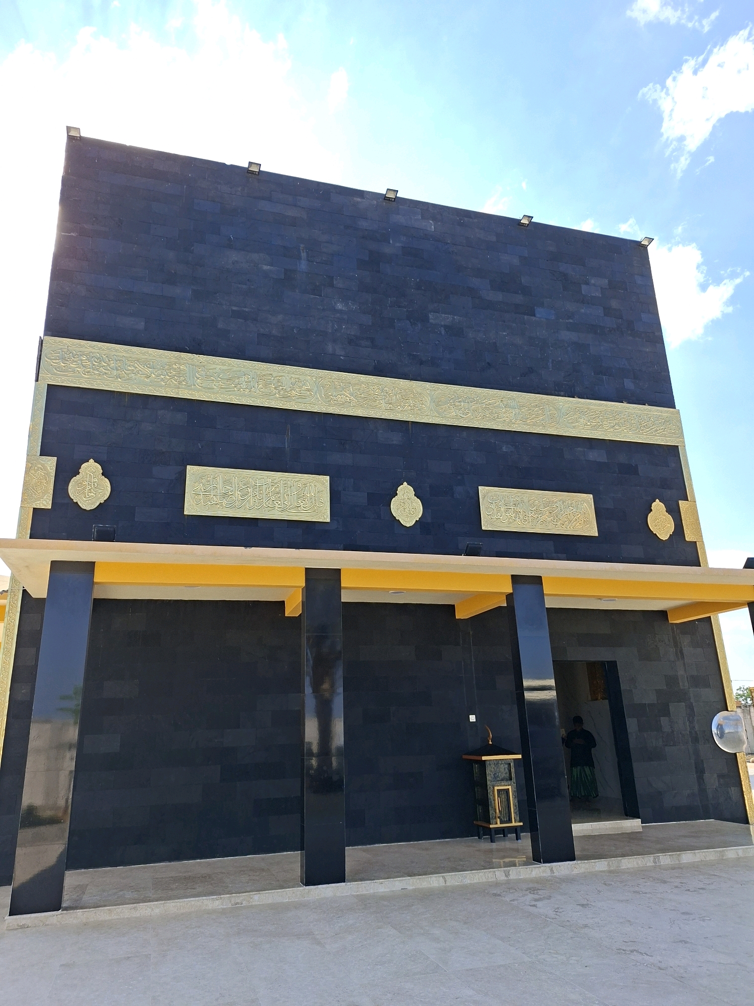 A low-angle, medium shot captures the exterior of a modern building resembling the Kaaba, located in Jrengik, Indonesia, under a bright blue sky with scattered clouds. The building is primarily clad in black stone tiles, with a prominent golden band featuring Arabic calligraphy running horizontally across its upper facade. Below this band, smaller gold decorative elements are affixed, with a rectangular inscription in the center. The lower portion of the building is also black stone, supported by large, black, square pillars. A yellow awning extends from beneath the upper facade, supported by these pillars. The ground in front of the building is paved with light-colored stone tiles. To the right of the entrance, a decorative lantern with gold trim and a black base sits on the ground. A person wearing a green sarong and a dark shirt is partially visible standing inside the building's entrance on the right side of the frame. The overall impression is one of religious architecture with a contemporary design.