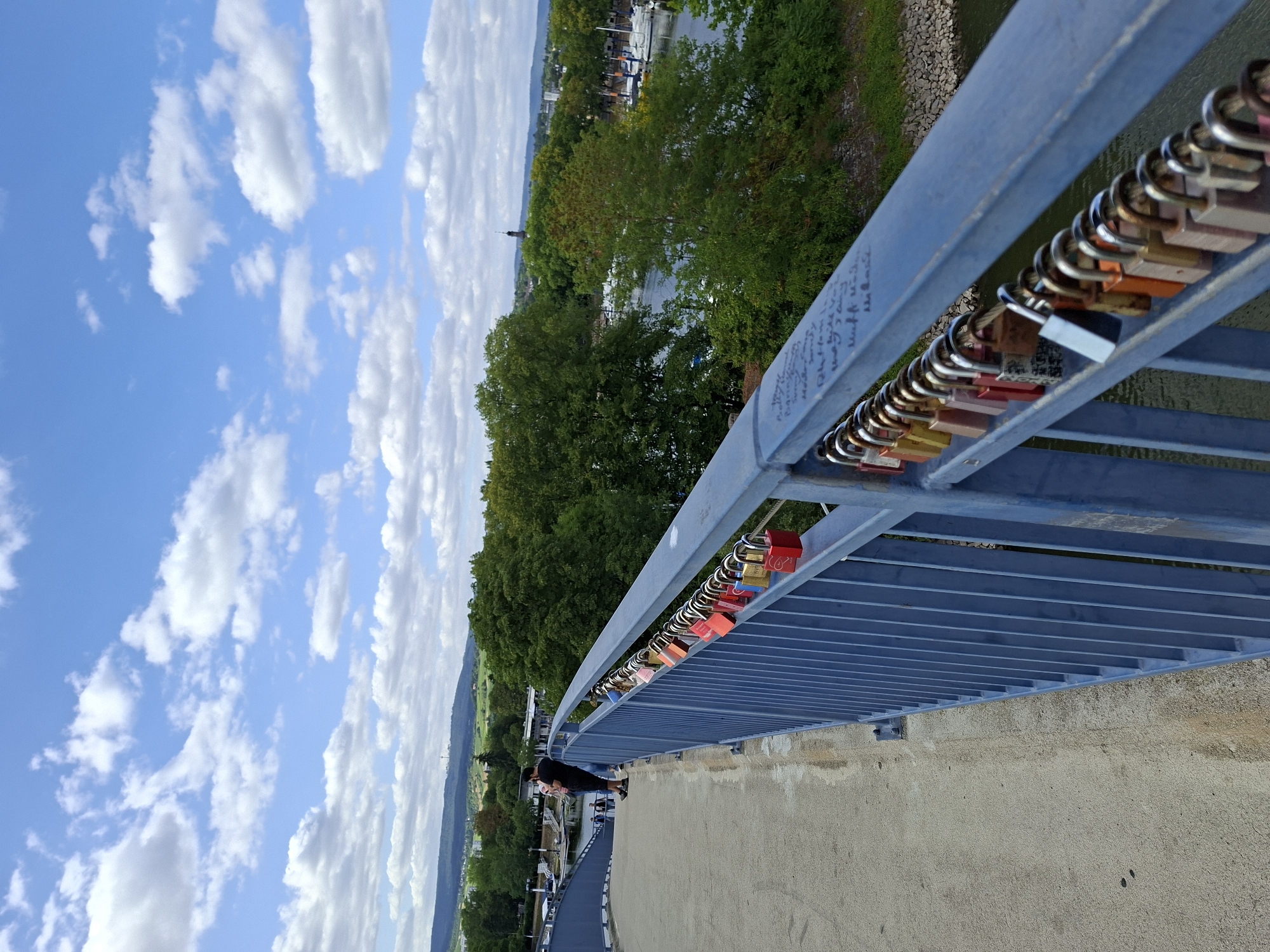 The image depicts a close-up view of a blue metal railing on a bridge, adorned with numerous colorful love locks. These locks, in shades of red, yellow, orange, pink, and silver, are attached to the railing, forming a continuous line. Some of the locks appear newer and shinier, while others are older and weathered. In the background, a body of water is visible, lined with lush green trees. Beyond the trees, the cityscape of Wiesbaden, Germany, unfolds, featuring buildings and a distant tower. A few people can be seen walking on the bridge in the distance, and the sky is a bright blue with scattered white clouds. The railing also has some handwritten inscriptions on it, likely names or messages.