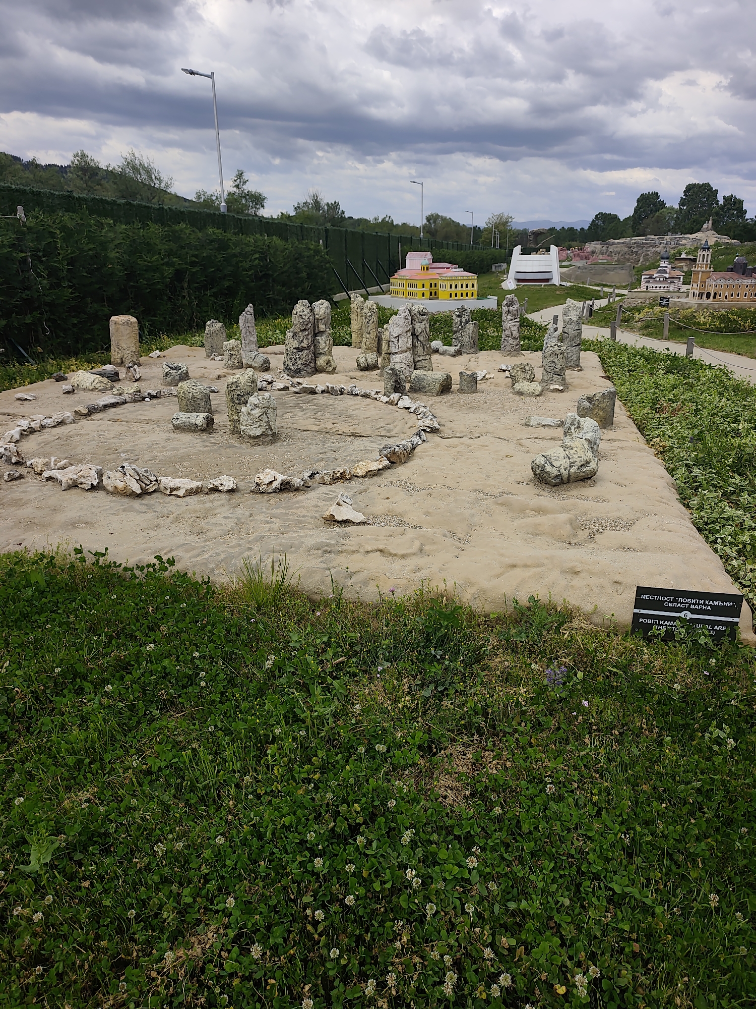 The image shows a miniature display of the "Pobiti Kamani" rock formations, located in Varna Province. The display consists of a circular arrangement of upright stones and a ring of smaller rocks. The miniature is set on a concrete platform surrounded by green vegetation. In the background, other miniature buildings are visible, as well as trees and an overcast sky.