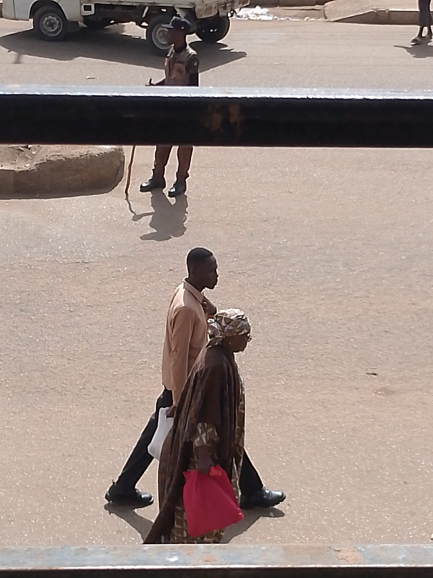 In Bukuru, Nigeria, a man in a tan shirt and black pants, accompanied by a woman in a patterned headscarf and a brown patterned dress carrying a bright red bag, walks across a paved surface. The woman is also wearing glasses and a long-sleeved garment. Behind them, another man in a camouflage uniform, a dark cap, and dark boots stands with a stick or cane. He appears to be on patrol. In the background, a white utility truck is parked on the right side of the frame. The scene takes place outdoors on a bright day, as indicated by the strong shadows cast by the subjects and the truck. The paved surface suggests a street or an open area. The overall impression is that of a casual pedestrian scene with a security presence. No visible text is present in the image.
