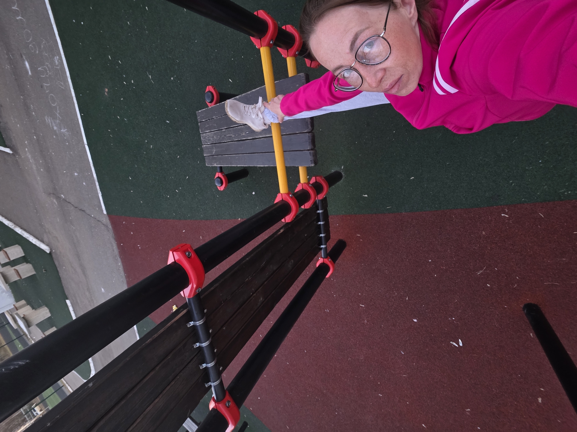 A woman is standing on a playground structure in Chaikovsky, Russia. She is wearing glasses and a pink jacket with white stripes. She has one foot on a wooden plank. The structure consists of black poles connected by red joints and yellow poles. The playground surface is multi-colored, with red, green, and possibly other colors. In the background, there is a gray surface with white markings.