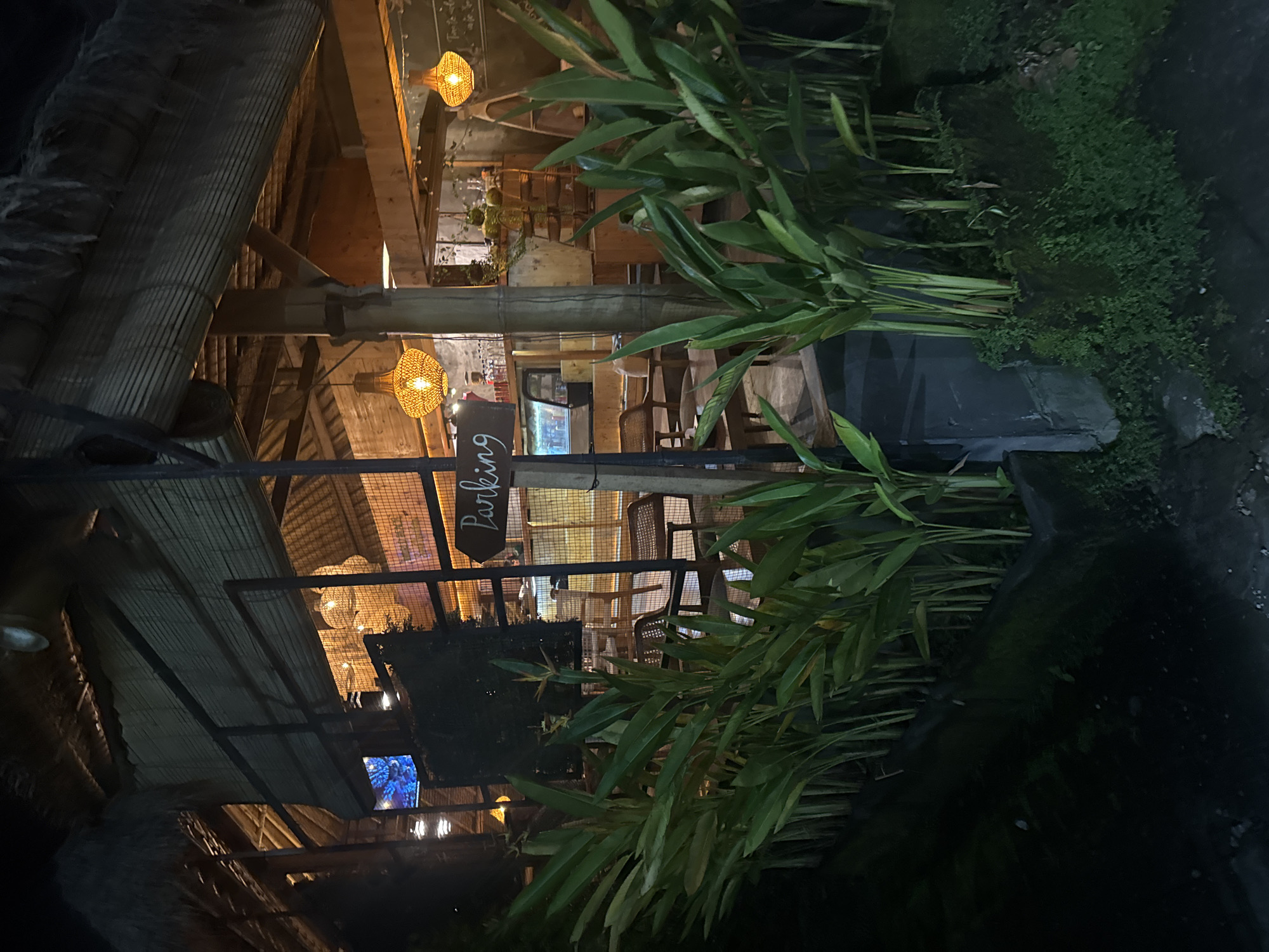 This image depicts an outdoor seating area of a restaurant or cafe in Ubud District, Indonesia, at night. The setting appears rustic and tropical, with a thatched roof supported by wooden beams and columns. Woven pendant lights illuminate the space, casting a warm glow. Lush green foliage, including large-leafed plants and dense greenery, frames the scene, suggesting a natural and tranquil environment.
In the foreground, a wooden sign with white lettering reads "Parking," indicating the direction to the parking area. Beyond the sign, visible through a metal fence, are rows of empty wooden chairs and tables, suggesting the establishment is either closed or not yet busy. The interior of the building shows more wooden structures and some artificial lighting, along with a television screen displaying a colorful image.
The time of day is clearly nighttime, with the artificial lights being the primary source of illumination. The overall mood is peaceful and inviting, characteristic of a relaxing evening dining experience. The location context of Ubud, known for its natural beauty and cultural heritage, is consistent with the tropical foliage and rustic architecture. There are no people actively engaged in any activity in this shot, emphasizing the quiet ambiance.