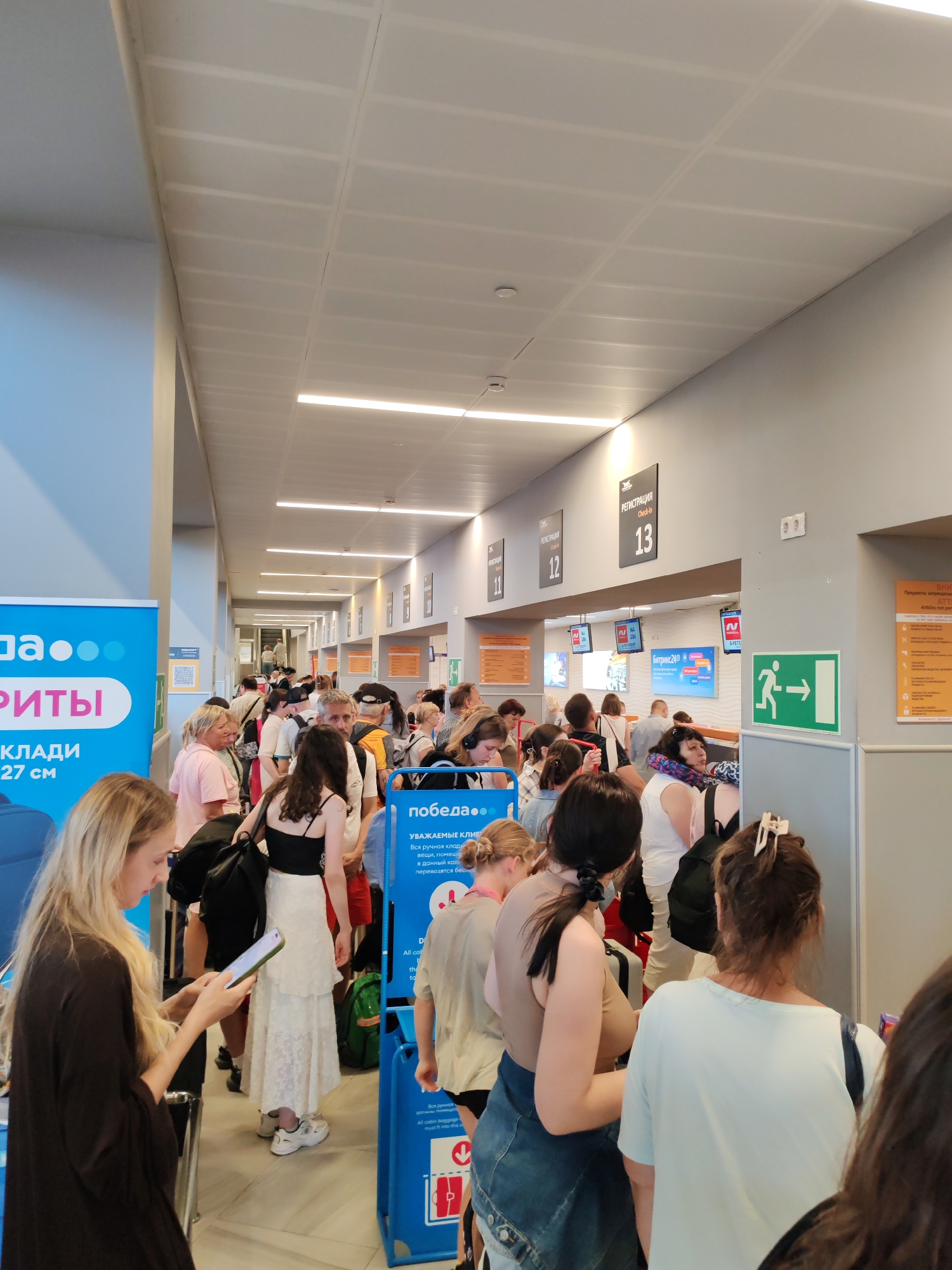 This is a vertical, eye-level, full shot of a crowded indoor scene, likely an airport or train station, given the presence of check-in counters and signage. A long queue of people, predominantly women, are lined up, suggesting they are waiting for a service, possibly check-in or boarding. Many are dressed casually, some with luggage, further indicating travel. The overall atmosphere appears somewhat somber or indifferent, with most individuals looking ahead or down.
The setting is a long, well-lit corridor with a dropped ceiling featuring fluorescent lighting. The walls are a neutral gray, with prominent signs indicating "РЕГИСТРАЦИЯ" (Registration) and numbers 11, 12, and 13 above different counters. A green directional sign with a running figure and an arrow pointing to the right is visible, indicating an exit or a specific path.
Visible text includes "РИТЫ" (likely part of a word related to offerings or services), "КЛАДИ 27 см" (possibly baggage dimensions), and "победа..." (which could be a brand name or airline). Other text includes "УВАЖАЕМЫЕ КЛИЕНТЫ" (Dear clients), "Вся ручная кладь" (All hand luggage), and information about dimensions and allowances, further confirming a travel-related context. One woman in the foreground is engrossed in her phone, while others in the queue appear patient. The time of day is not explicitly discernible but the bright lighting suggests it's daytime. The location is identified as Khrabrovo, Russia.