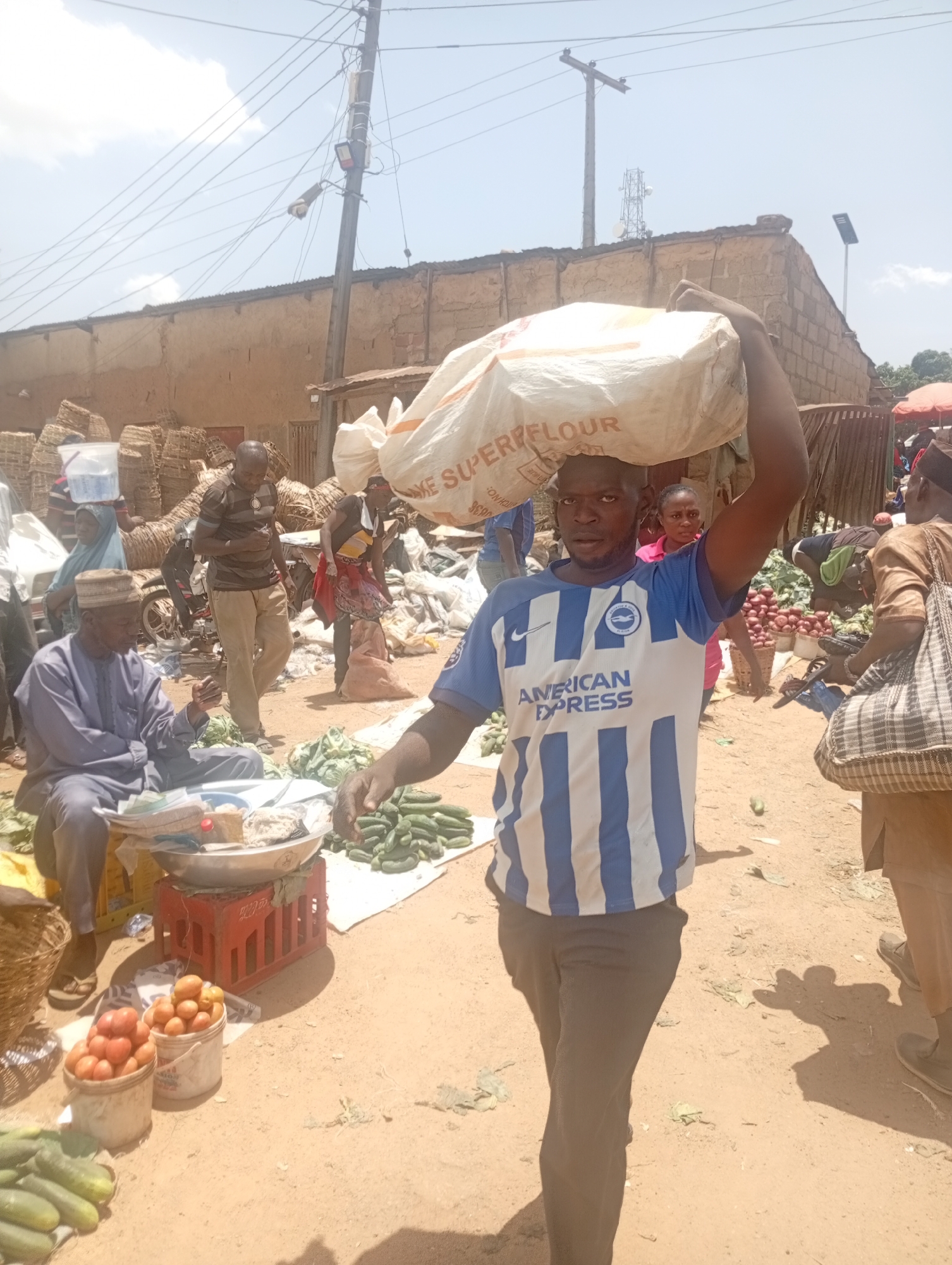 A man in a blue and white striped shirt is carrying a large white bag on his head, walking towards the viewer. The bag is labeled "SOME SUPERFLOUR." He is surrounded by a busy outdoor market scene with several people. To his left, an older man in a light blue robe sits next to a scale, and there are tomatoes and cucumbers nearby. Other people stand around, some looking at goods or talking to each other. Woven baskets are stacked in the background. The setting appears to be a crowded marketplace, likely in a tropical or subtropical climate, on a bright, sunny day.