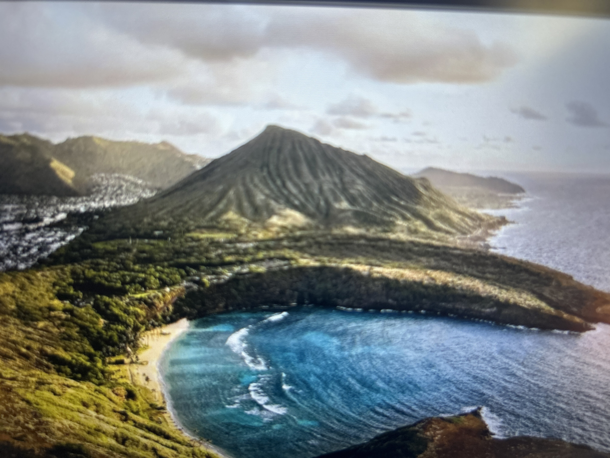 The image shows an aerial view of a coastal landscape. A prominent mountain dominates the scene, featuring a striated surface suggesting volcanic or geological formations. Lush green vegetation covers the base of the mountain and the surrounding landscape. A bay with turquoise water is visible in the foreground, with white waves breaking along the shore. A sandy beach is situated along part of the bay. The sky is overcast with clouds, providing soft lighting. In the background, a city or town is partially visible along the coastline. Given the lack of specific landmarks recognizable as being in Varna, Bulgaria, definitive identification is not possible.