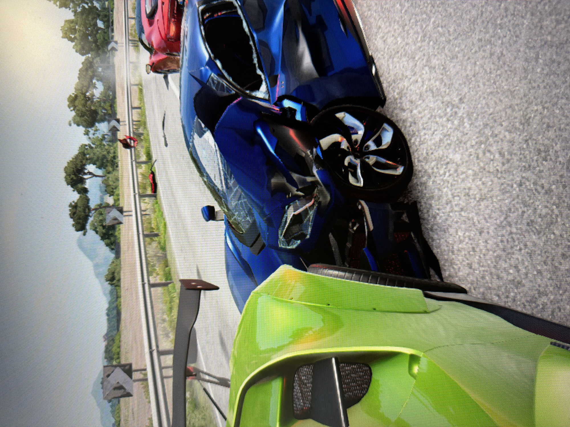 This media file is a photograph of a screen displaying a scene from what appears to be a racing video game, evidenced by the distinct pixelation. The image is oriented vertically (rotated 90 degrees counter-clockwise).
The central focus is on three sports cars on an asphalt road. In the immediate foreground, to the bottom right of the image (when viewed upright), is the rear section of a bright lime-green sports car, possibly a Lamborghini, featuring a prominent black rear spoiler and mesh grilles.
Dominating the midground is a metallic blue sports car, heavily damaged on its front-right side, with the wheel clearly visible and mangled bodywork. Its angle suggests it may have been involved in a collision or is static after one. Behind it, partially visible and also appearing distorted or damaged, is a red sports car.
In the background, along the left side of the road, a person wearing red clothing can be seen in the distance, standing near what looks like a guardrail or barrier. The road extends into the distance, flanked by lush green trees and hilly or mountainous terrain under a bright, possibly hazy sky. Road signs indicating a curve are visible along the roadside. The overall impression is a moment captured after a high-speed incident on a scenic route or race track during daytime. No discernible text is visible in the image.