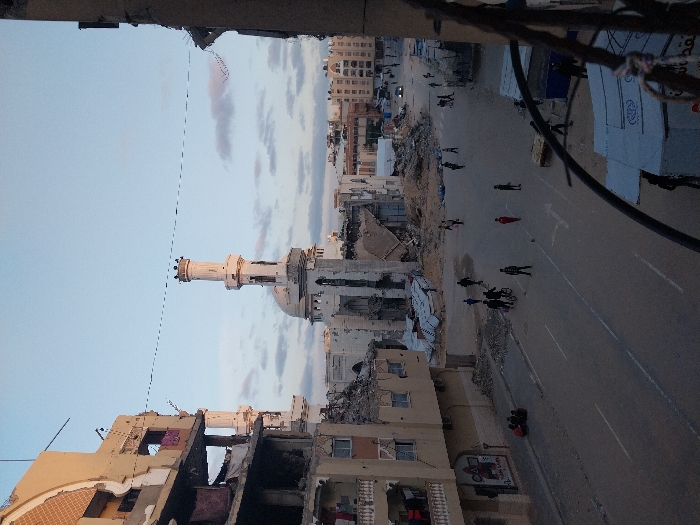 The image captures a street scene in Khan Yunis, Palestinian Territory, under natural daylight with a mostly clear, pale blue sky and scattered white clouds. A prominent white mosque, featuring a tall minaret with a crenellated top and a large dome, stands in the mid-ground, appearing partially damaged and surrounded by significant piles of rubble and debris. Flanking the paved street are multi-story buildings exhibiting extensive structural damage, including collapsed sections, exposed interiors, and missing facades.
On the left side, a multi-story building, heavily damaged but partially intact, shows clothes hanging from balconies on its upper floors. Below it, on the sidewalk, a small group of approximately three to four individuals are seated. On the right side, a severely damaged building forms part of the foreground, its structural elements partially obscuring the view.
The paved street, marked with white lines indicating traffic lanes and possibly pedestrian crossings, is populated by numerous individuals. In the mid-ground, a group of people are walking and standing; one person is seen riding a bicycle with a passenger. More distant figures are visible further down the street, where additional damaged structures and rubble are present. Temporary, tent-like shelters are discernible on the right side of the street in the foreground. A utility line spans horizontally across the upper mid-portion of the sky.
