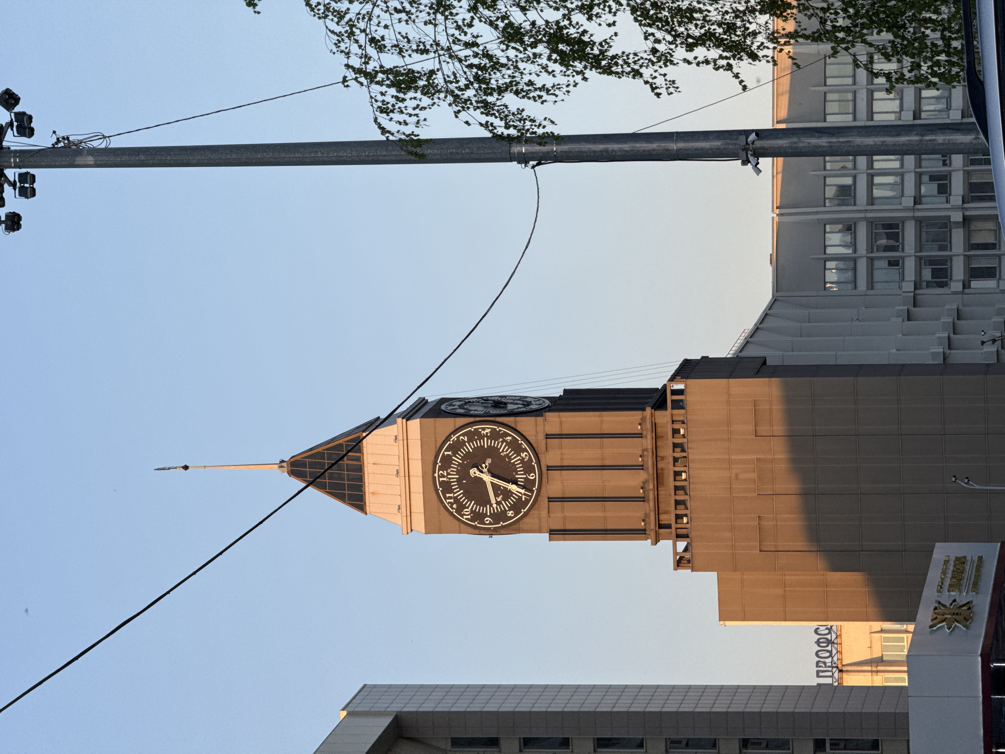 The image shows the Krasnoyarsk Clock Tower in Russia from a ground-level perspective. The clock face displays a time between 10:10 and 10:15. A lamppost with lights is visible in the upper left, and electrical wires run across the sky. The surrounding buildings are a mix of modern and older architectural styles. Trees are partially visible at the top of the frame.