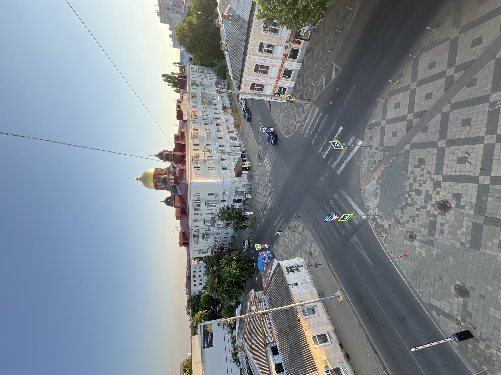 This image captures an aerial view of a city intersection in Krasnodar, Russia, bathed in the warm light of a setting sun. Dominating the skyline is a prominent building with a golden dome and red roof, reminiscent of a church or cathedral. The intersection itself is paved with cobblestones and asphalt, featuring pedestrian crosswalks and traffic signs. Several cars are visible on the roads, including a blue sedan, a white car, and two dark-colored vehicles. Adjacent to the intersection are various buildings, including a multi-story apartment complex, smaller, older structures with gray roofs, and a building with a sign that reads "ГОЛДАВСКИЙ ДОМ" (Goldavsky House). Trees line some of the streets, adding a touch of greenery to the urban landscape. The sky is a clear, pale blue, with a few faint clouds visible. The overall scene suggests a peaceful, late afternoon or early evening in a European-style city.