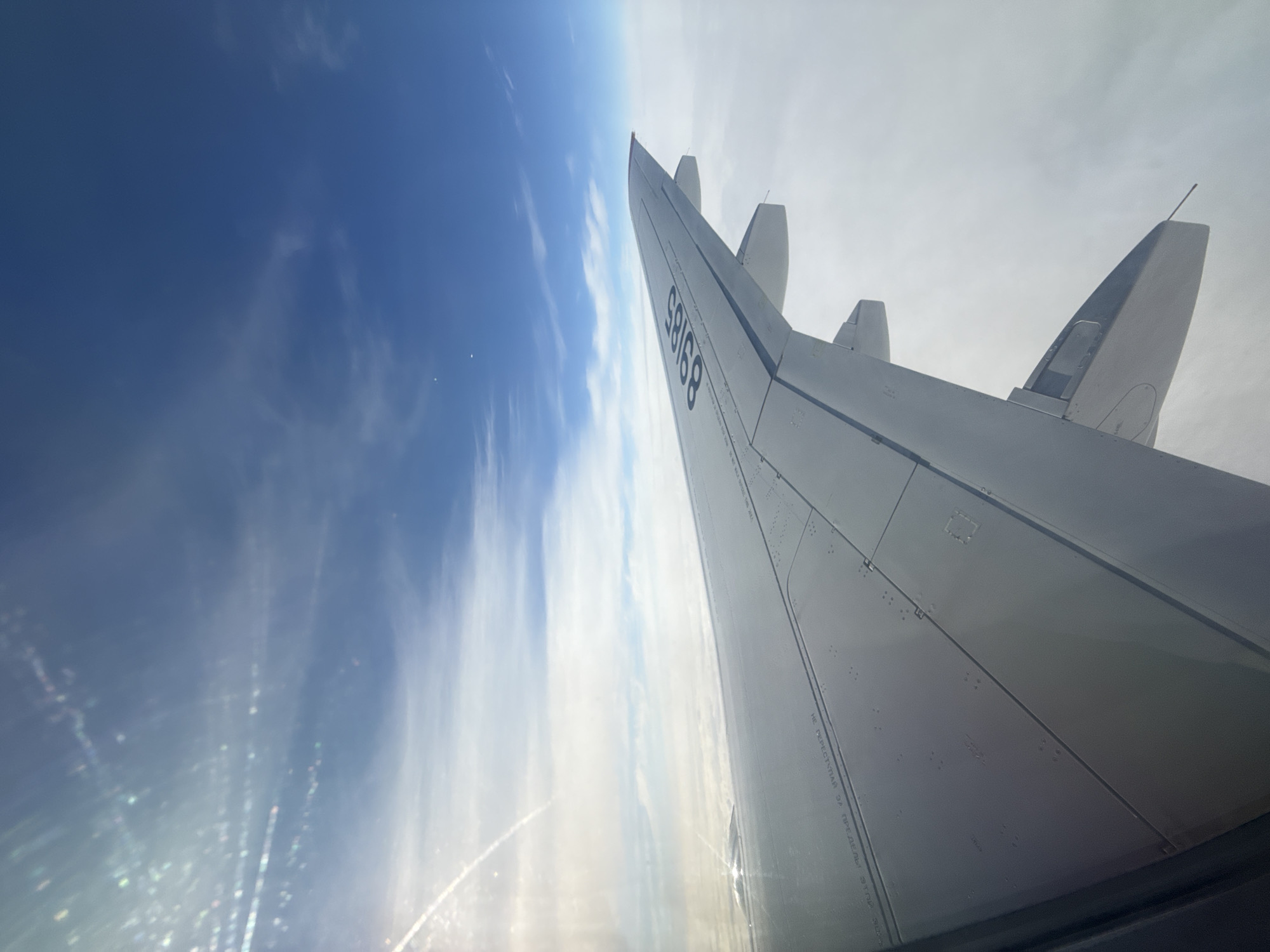 A photograph taken from inside an aircraft depicts an airplane wing and sky. The wing is white and angled upwards, with the number "88189" visible on its surface. The sky is a mix of blue and white, with some cloud formations. The photograph's orientation is slightly tilted. The location is not discernable from the image itself. Новое Мартьяново, Russia is not discernible from the image itself.