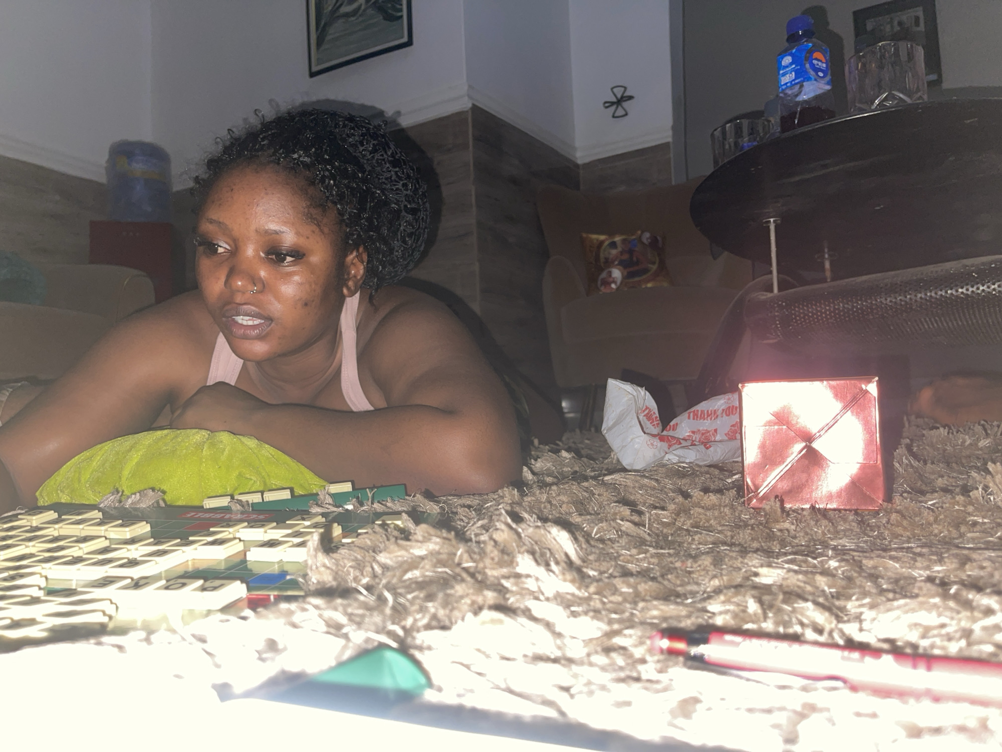 A young woman is depicted lying on her stomach on a shaggy brown rug indoors, deeply engrossed in a game of Scrabble. She is positioned at the lower left of the frame, propped up on her forearms with a lime green pillow supporting her chest. She wears a light pink sleeveless top and has dark, curly hair. Her gaze is directed towards the Scrabble board in front of her, and her expression suggests concentration or contemplation. A small nose ring is visible on her left nostril.
The indoor setting appears to be a casually furnished room, possibly a living area, characterized by its warm, subdued lighting. In the immediate foreground, to the right, there is a shiny red, square-shaped object, possibly a small gift box, and a red pen or marker. A white plastic bag with the words "THANK YOU" printed on it rests on the rug. The Scrabble board itself clearly shows the word "SCRABBLE" and several letter tiles.
In the mid-ground and background, various household items are visible. A dark, circular table holds a blue water bottle, identifiable by the word "WATER" and other text on its label, and two clear glass tumblers. Further back, an armchair with a patterned cushion, a small red storage unit with a blue roll on top, and a framed piece of artwork adorn the walls. A decorative metal ornament is also affixed to a wall corner. The overall ambiance suggests a relaxed evening activity in Kuchingoro, Nigeria.