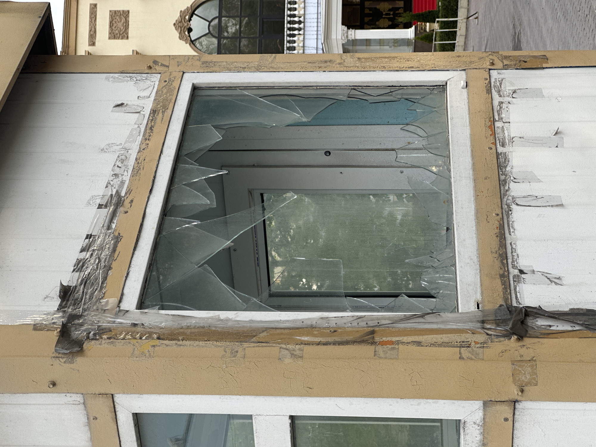 A damaged window is shown at an upward angle. The window glass is shattered, with shards remaining in the frame. The window frame appears to be white, set within a larger structure that is painted with a cream or light brown color. There is tape of varying types used to hold the broken pieces in place. The paint on the outer structure shows signs of peeling and wear.
The location is Moscow, Russia, based on visual elements such as architecture.