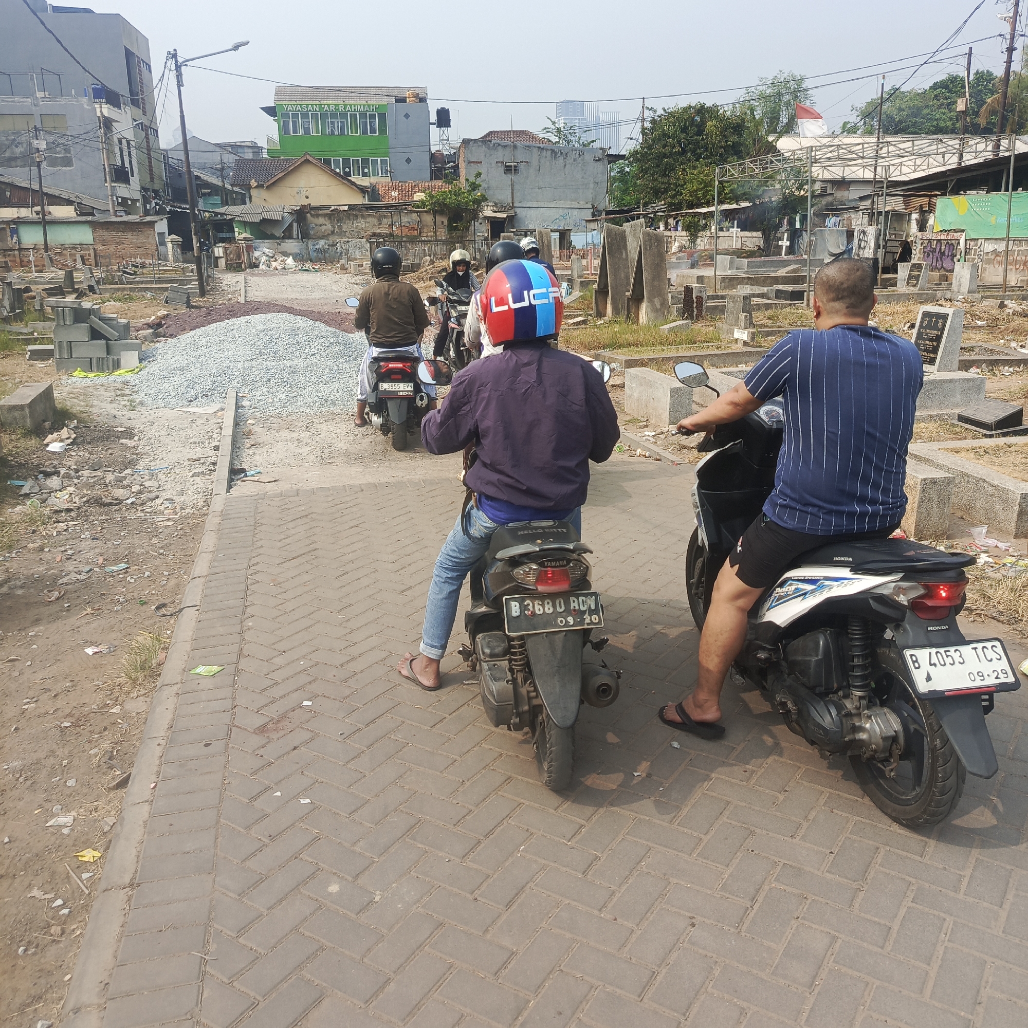 The image shows a street scene in South Jakarta, Indonesia, likely near a cemetery and construction area. In the foreground, three people are riding on motorcycles. The rider in the center is wearing a blue, red, and white helmet. The rider on the left is wearing a brown jacket and a black helmet. The rider on the right is wearing a blue and white striped shirt and black shorts. The motorcycles are parked on a brick-paved road next to a dirt path with piles of gravel. In the background, there are buildings, including a green building with the text "YAYASAN 'AR-RAHMAH'" and a cemetery with tombstones. The sky is overcast.