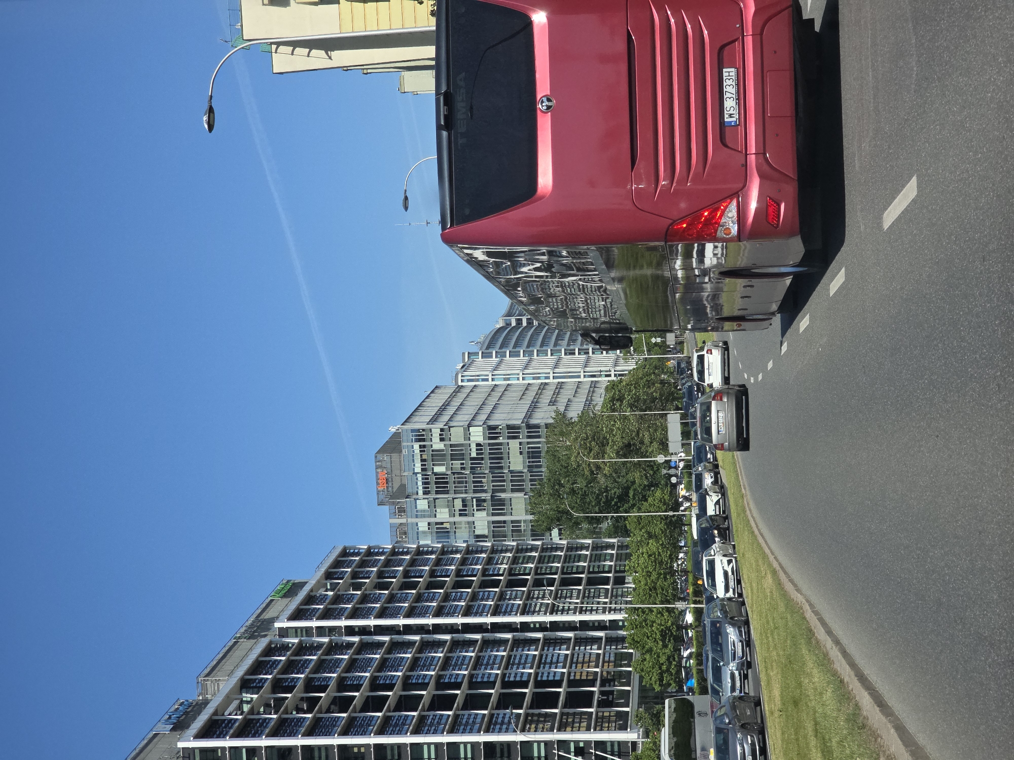 The image shows a street scene in Warsaw, Poland, with modern buildings lining the road. A red bus is prominently positioned in the right side of the frame, and several other vehicles are visible in a queue along the road, next to a patch of green grass. The sky is clear and blue. The building with the distinctive grid-like facade and the building behind it are located at Aleje Jerozolimskie in Warsaw.