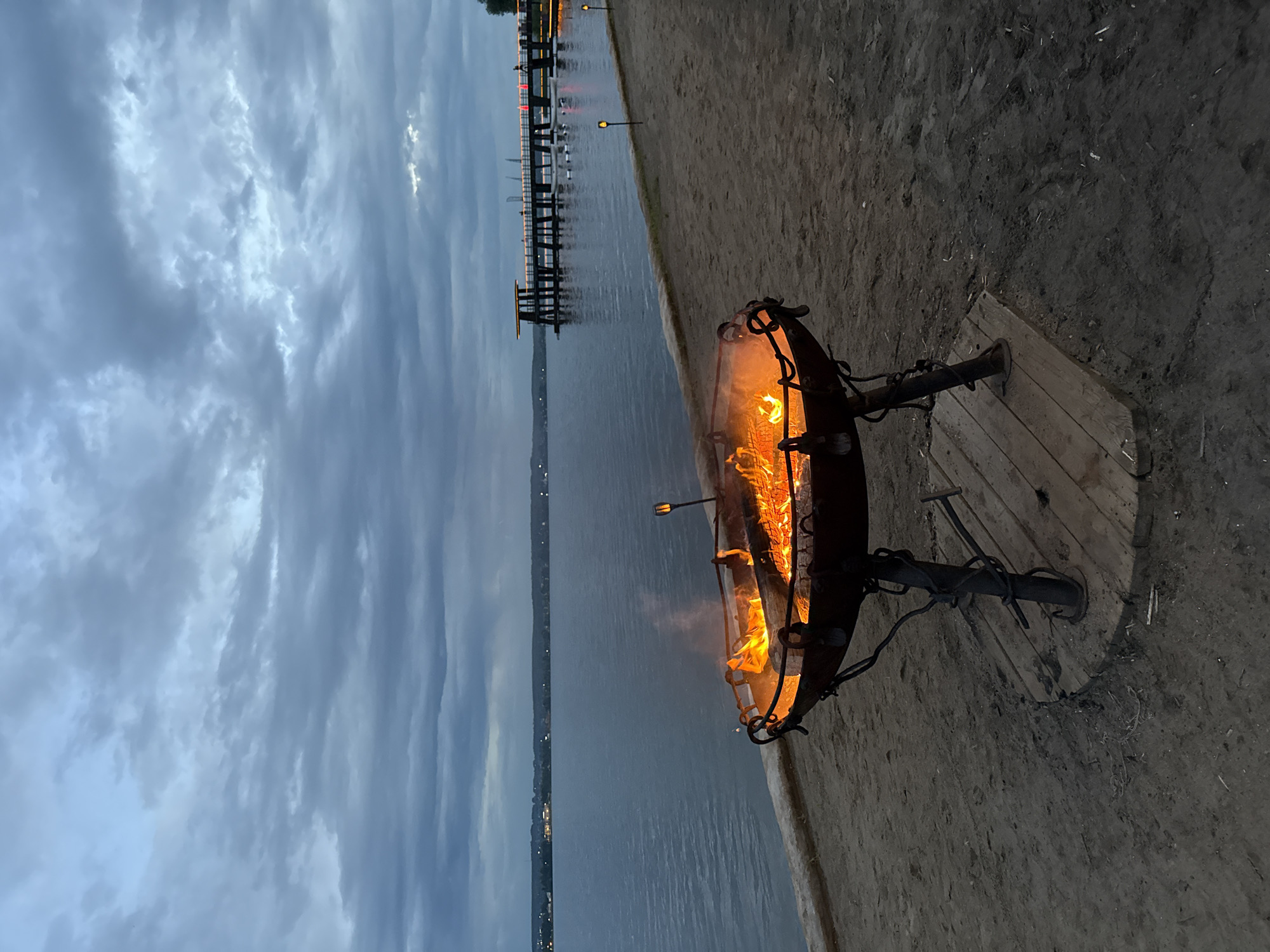 This is a medium shot, eye-level, outdoor photograph of a fire pit on a sandy beach at dusk. The fire pit, made of dark metal with ornate legs, is filled with burning logs and flames, casting an orange glow. The fire pit is situated on a wooden platform placed on the sand.
Behind the fire pit is a large body of water, likely a lake or sea, stretching to the horizon. The water is calm and reflects the twilight sky. In the distance, across the water, a silhouette of a city or town can be seen with faint lights. A long pier, illuminated with small lights, extends into the water on the right side of the frame. Several small, lit garden lights are also visible along the edge of the beach.
The sky is overcast with dark, dramatic clouds, suggesting either the end of a stormy day or an approaching storm. The overall mood of the image is serene and a little melancholic, with the warmth of the fire contrasting with the cool tones of the sky and water. The scene appears to be set in Votkinsk, Russia, specifically near the shore of Lake Votkinskoye.