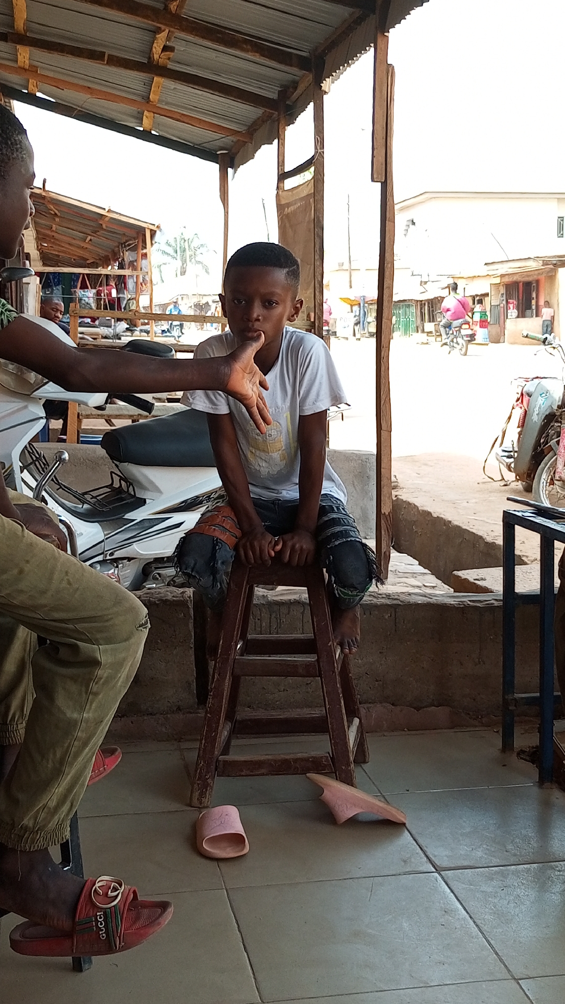 A young boy sits on a wooden stool inside a building with a corrugated metal roof, located in an unidentified urban area. He is fair-skinned, has short hair, and wears a white t-shirt and worn-out denim jeans with patches. His right hand is resting on his chin, and he is looking directly at the camera.
An adult with dark skin is partially visible on the left side of the frame. They are wearing a light-colored shirt and khaki pants, and one hand is extended towards the boy.
Behind the boy, there is a wall, and through an opening, a street scene is visible. There are buildings, people, and a motorcycle. In the foreground, there are two pairs of sandals on the tiled floor. One pair is red with a Gucci emblem and the other is pink. A motorcycle is parked next to the boy, as well.