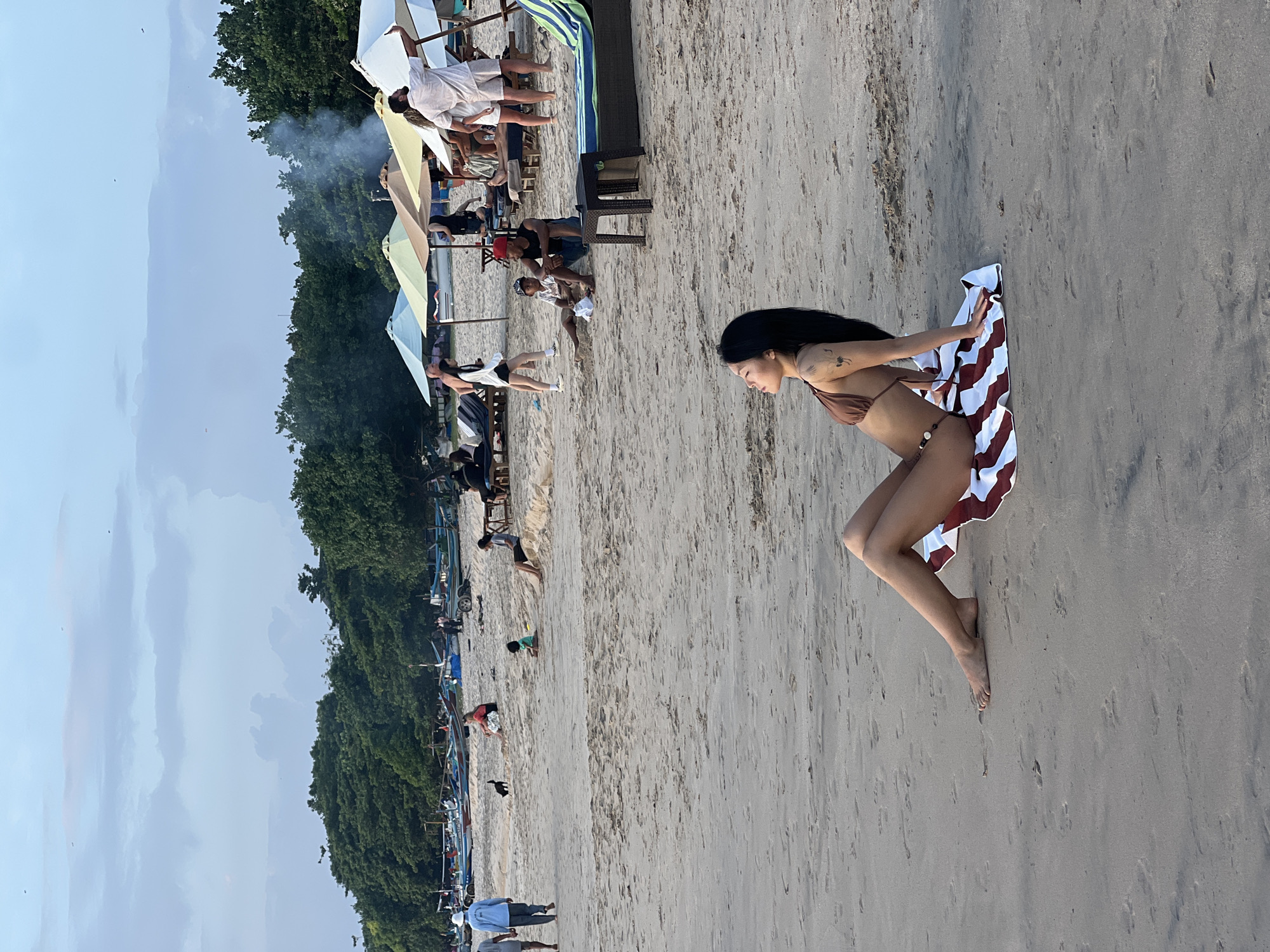 An image captures a beach scene in Kuta Selatan, Indonesia, featuring a young woman in a brown bikini sitting on a striped towel on the sand. Her body is angled to her left, with her head tilted back and eyes closed, seemingly enjoying the moment. In the background, several beach umbrellas, lounge chairs, and groups of people are visible, suggesting a popular tourist spot. The shoreline is lined with numerous traditional Indonesian fishing boats, characteristic of the region. The sky above is a muted blue with scattered clouds, and a faint haze or smoke is present in the air, possibly from a distant barbecue or cooking fire. The overall atmosphere is relaxed and casual, typical of a beach setting.