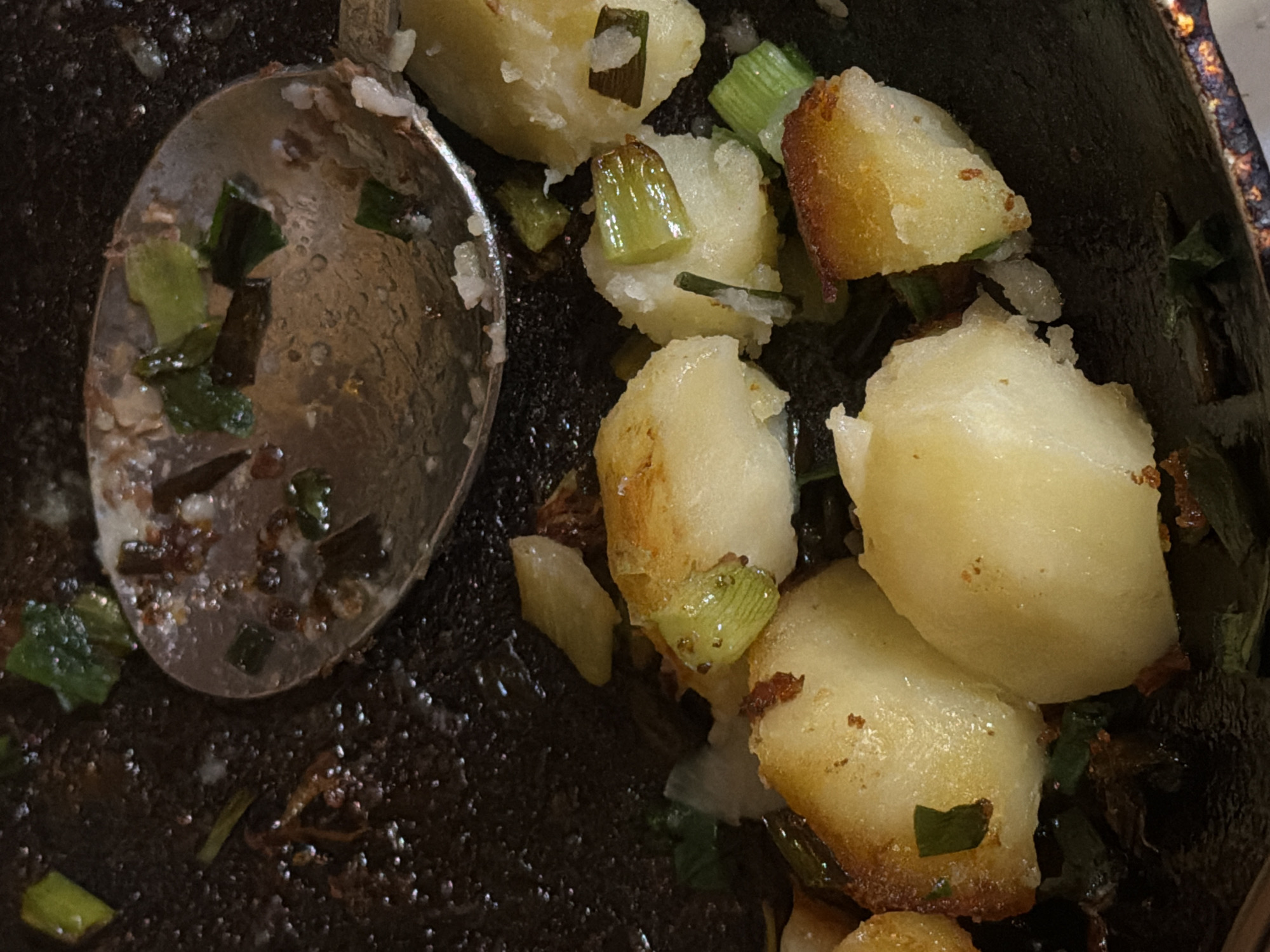A close-up, overhead shot shows roasted potatoes with scallions in a pan, as if recently served or prepared. The pan is dark and appears to be cast iron, with some residue and browning. A silver spoon rests in the upper portion of the pan, partially scooping some of the potatoes and green onions. The potatoes are roughly cut into bite-sized pieces, some with golden-brown edges from roasting, while others remain pale yellow. The green onions are chopped into short segments and interspersed with the potatoes. The overall impression is of a rustic, home-cooked meal. This image is not associated with Satino-Russkoe, Russia, as there is no visual information to support such a location.