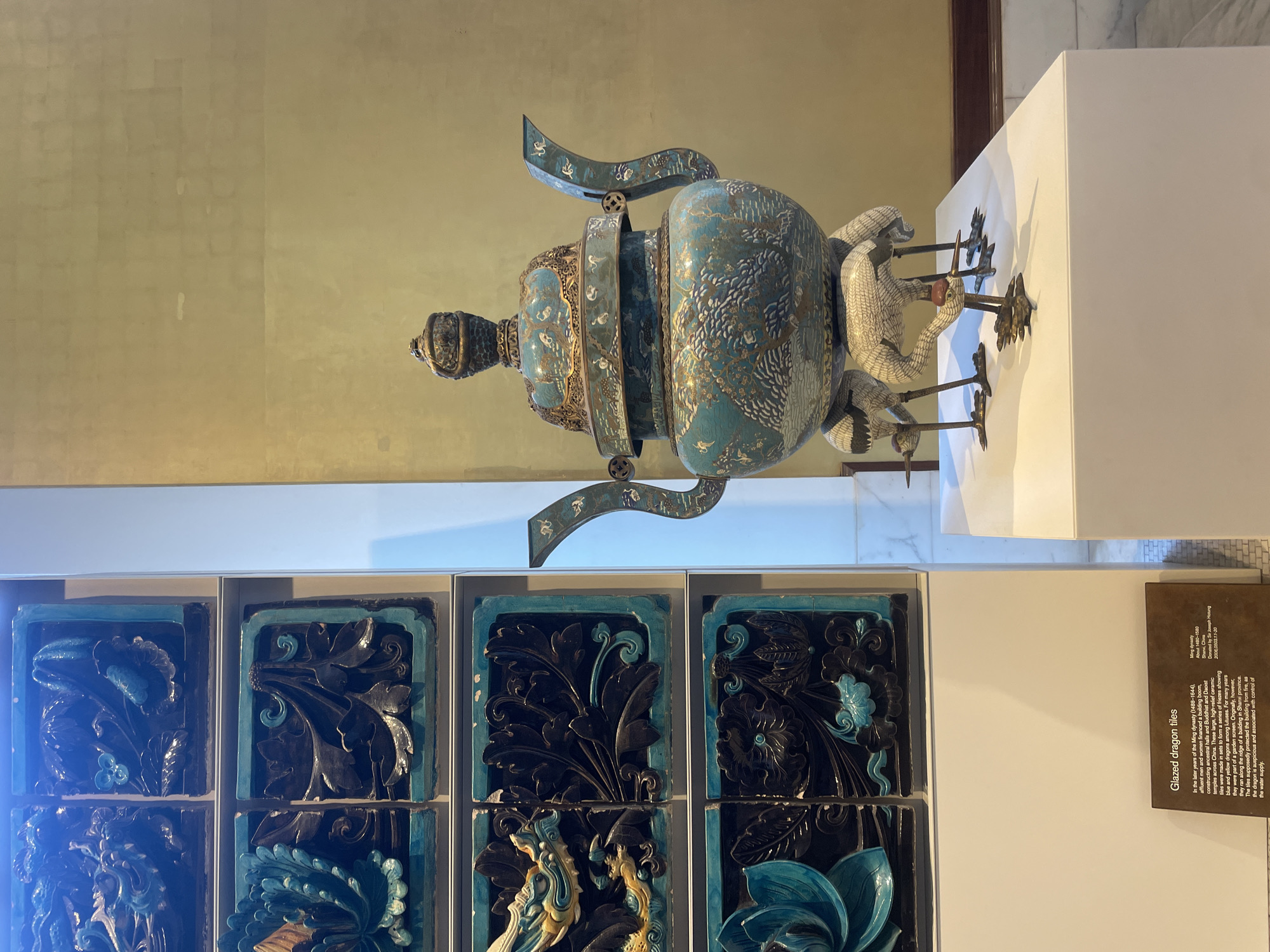 The media file showcases a display of ancient Chinese glazed dragon tiles, likely in a museum setting. The tiles, featuring intricate relief carvings of dragons and lotus flowers in shades of blue and black, are arranged in a grid on a shelf. A descriptive plaque beside the display reads: "Glazed dragon tiles" and provides information about their origin, materials, and historical context, noting they are from the Ming dynasty (about 1480-1580) and from Shan, China. Above the tiles, a large, ornate ancient Chinese vessel, possibly a censer or urn, is displayed. This vessel is primarily teal in color with intricate gold detailing and decorative handles. It is positioned on a white pedestal, with a pale yellow wall in the background. The overall scene suggests an exhibition of historical artifacts, specifically focusing on Chinese art and craftsmanship. The lighting appears to be indoor museum lighting, casting shadows and highlighting the textures of the objects.
