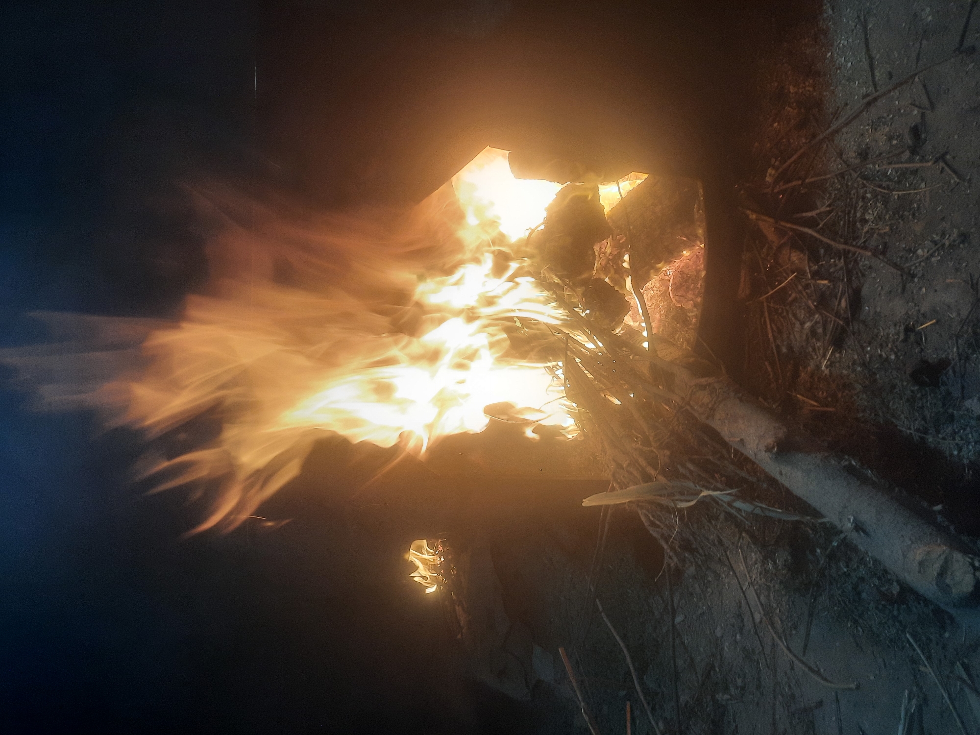 A nighttime scene depicts two fires burning in the city of Vosiobod, Tajikistan. The primary fire, larger and more intense, is contained within an open-sided structure, likely a metal brazier or kiln. Flames, ranging in color from bright yellow to orange, erupt from the burning materials, which appear to be wood and other combustible debris. The fire illuminates the immediate surroundings with a warm glow, casting shadows and highlighting the rough texture of the ground, which is covered in dirt, small stones, and scattered twigs and branches. In the background to the left, a second, smaller fire burns with less intensity, appearing to be a more modest bonfire. Both fires are generating smoke, which drifts upwards and into the dark night sky, creating a hazy atmosphere. The overall impression is one of a rural or semi-urban environment at night, where fires are being used for warmth or some other purpose.