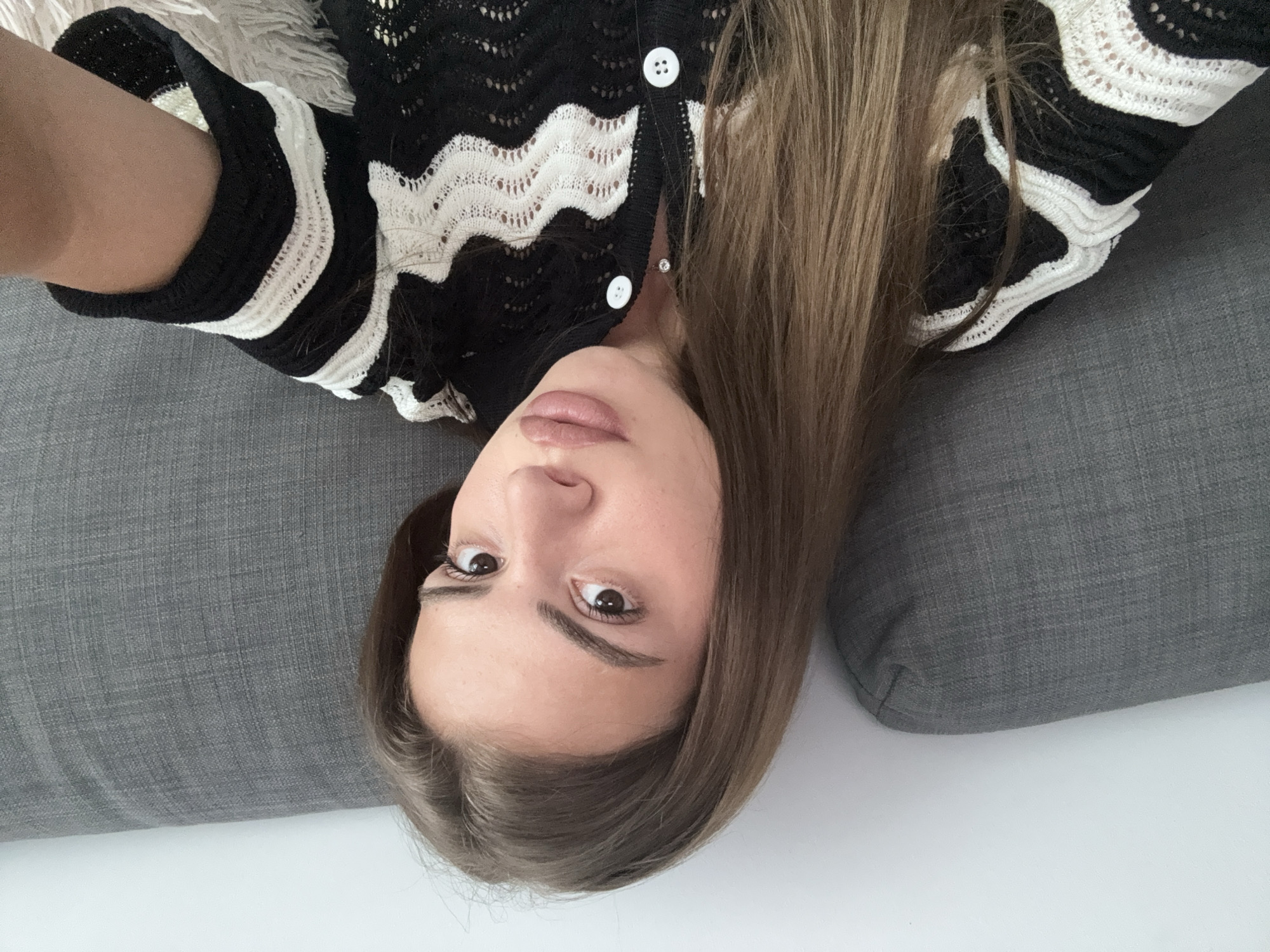 The image captures a young woman lying down, looking directly into the camera. The photo is oriented such that her head is towards the bottom, and her body extends upwards, angled to the right. She has long, straight, light brown hair that cascades around her head. Her eyes are open, dark, and focused on the viewer, with a neutral to slightly pensive expression. Her lips are naturally colored and slightly parted.
She is wearing a black and white knitted cardigan or top, featuring a distinct wavy or zigzag pattern. The white sections of the garment have an openwork, lacy knit texture, contrasting with the solid black areas. Small, round white buttons fasten the front of her cardigan. A tiny, clear stud earring is visible in her right ear.
She is resting on a grey, textured upholstered surface, likely a sofa or cushions, which forms the main background. A soft, white, fluffy material, possibly a blanket or pillow, is visible near her left shoulder. The lighting suggests an indoor setting, with no specific time of day or location cues. No text is visible in the image.