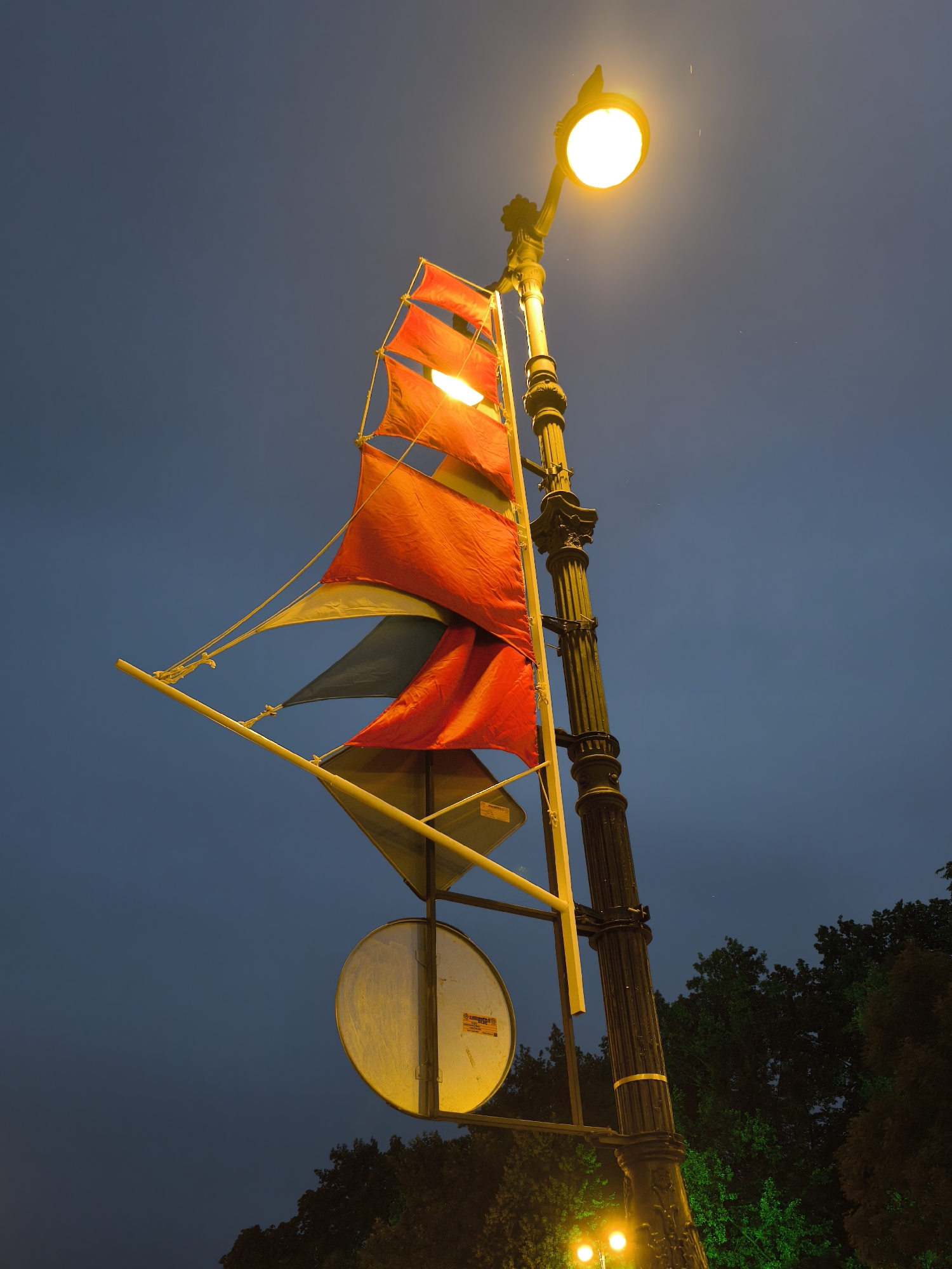 A street lamp with an illuminated bulb shines against a dark blue sky in Saint Petersburg, Russia. The lamp post is adorned with decorative elements and has a black, ribbed column. Attached to the lamp post is a structure with several colorful fabric "sails" in red, yellow, and gray, resembling a ship's rigging. Below the sails, a circular, reflective sign is visible, illuminated by the street lamp. In the background, dark green trees are partially lit by green light, and two smaller illuminated bulbs can be seen near the bottom of the image. The sky appears overcast with very faint raindrops visible.