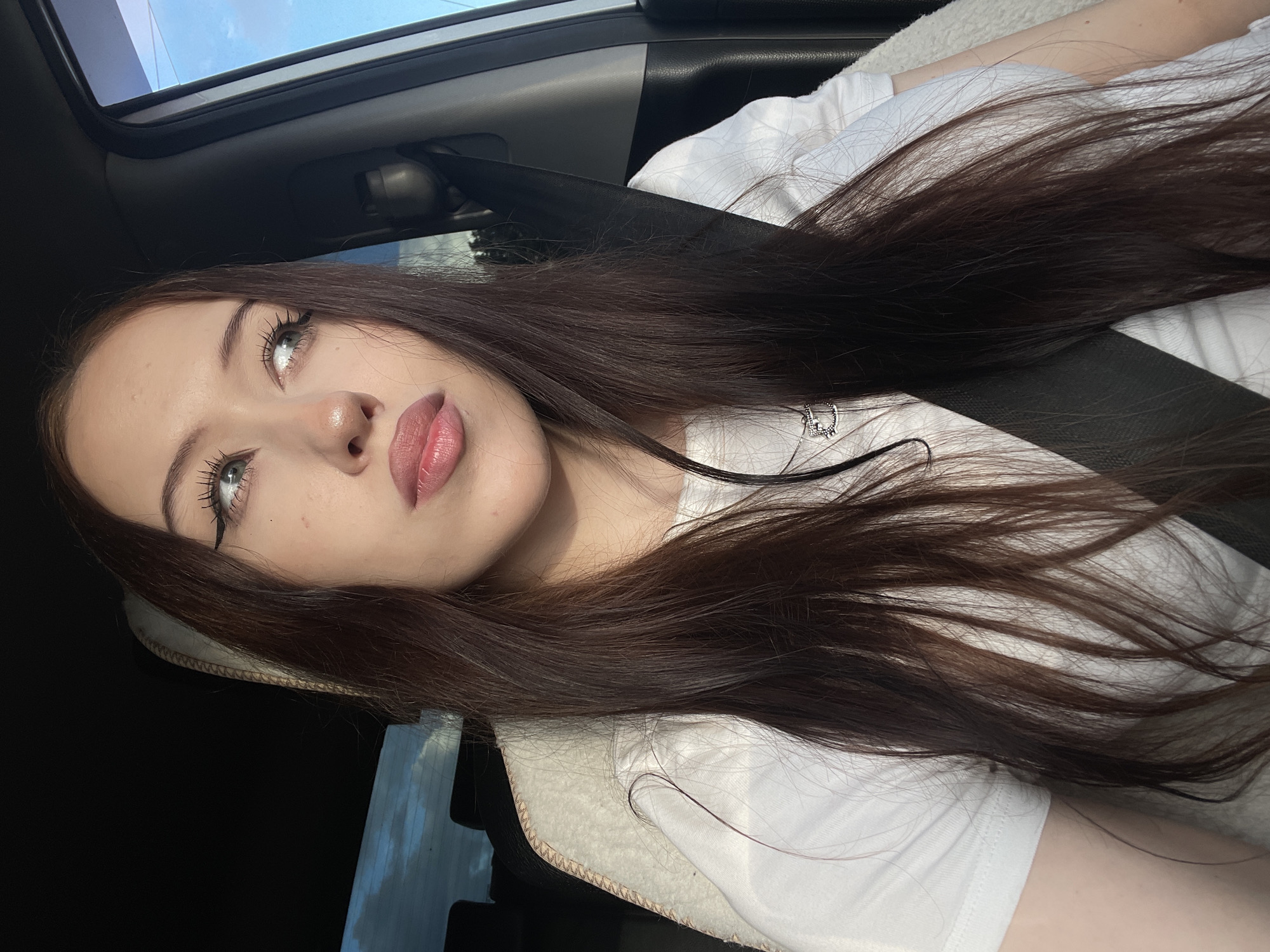A young woman with long, dark brown hair is captured in a selfie taken inside a car. She is wearing a white t-shirt and a silver necklace with a small charm. Her makeup is prominent, featuring winged eyeliner and lip gloss. She is looking upwards and slightly to the left, with a relaxed expression. The car's sunroof is open, revealing a bright blue sky with some clouds, suggesting it is daytime with fair weather. The interior of the car is visible, including a black seatbelt and a white headrest or cushion. The overall lighting indicates it's likely a sunny day. The image appears to be taken in Gavrilovka, Russia. No text is visible in the media file.