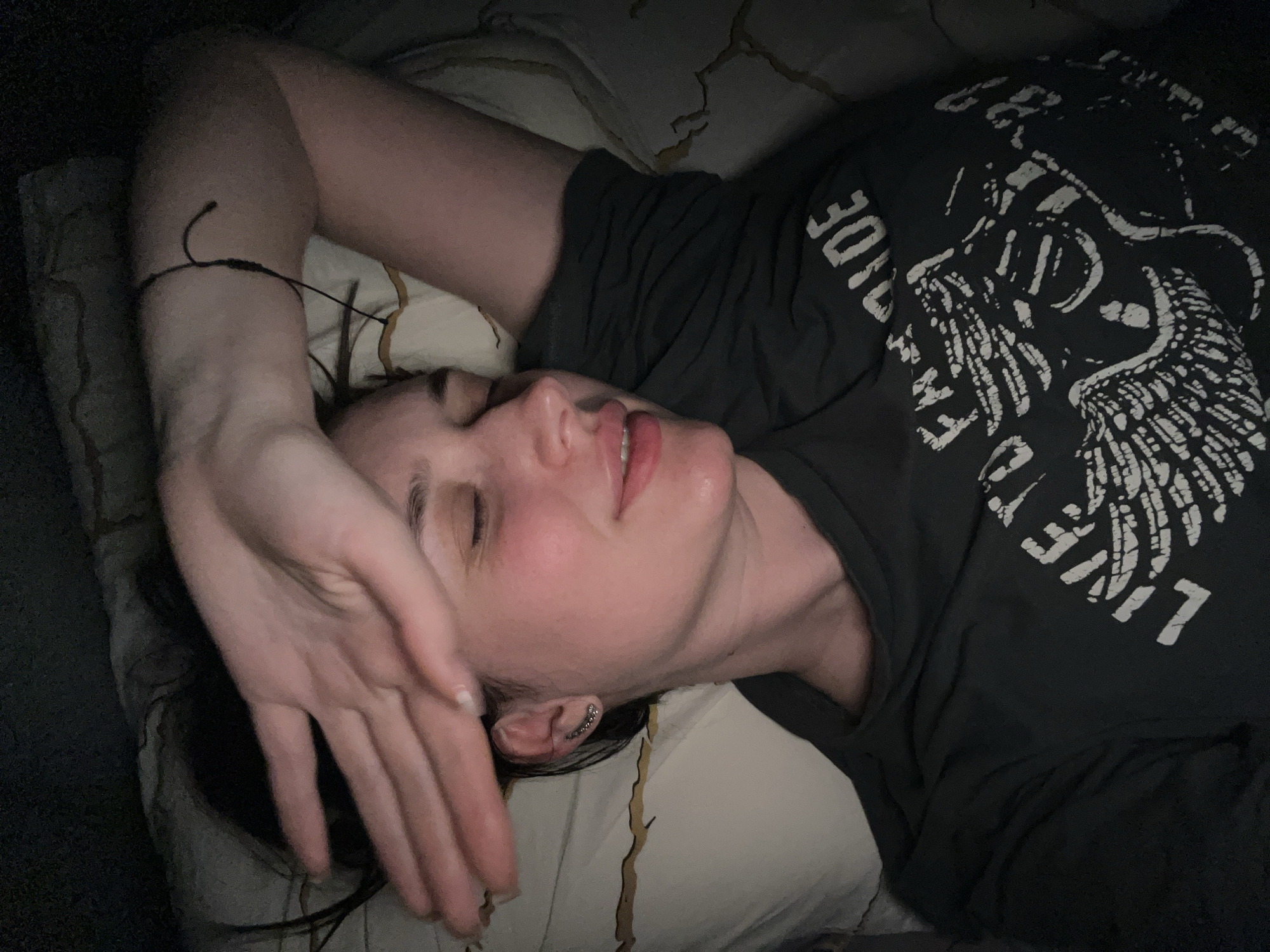 A person is depicted lying on their back in an indoor, dimly lit environment, likely a bed. Their head is turned slightly to the viewer's left, with eyes closed or partially closed and lips slightly parted. The person's right arm is bent, with the right hand resting on their forehead and upper face. They wear a dark-colored short-sleeved t-shirt featuring a white graphic design, which includes text such as "LIVE TO RIDE" and an emblem resembling a winged skull or creature. The visible left ear has a silver-colored ear cuff or multiple small earrings along the helix. A thin, dark cord-like item, possibly a bracelet, is present on the left wrist. Dark hair is visible against a light-colored pillow with subtle patterns. The bedding includes a pillow and a patterned sheet or blanket. The image contains no discernible features that can identify its location as Tyumen, Russia.