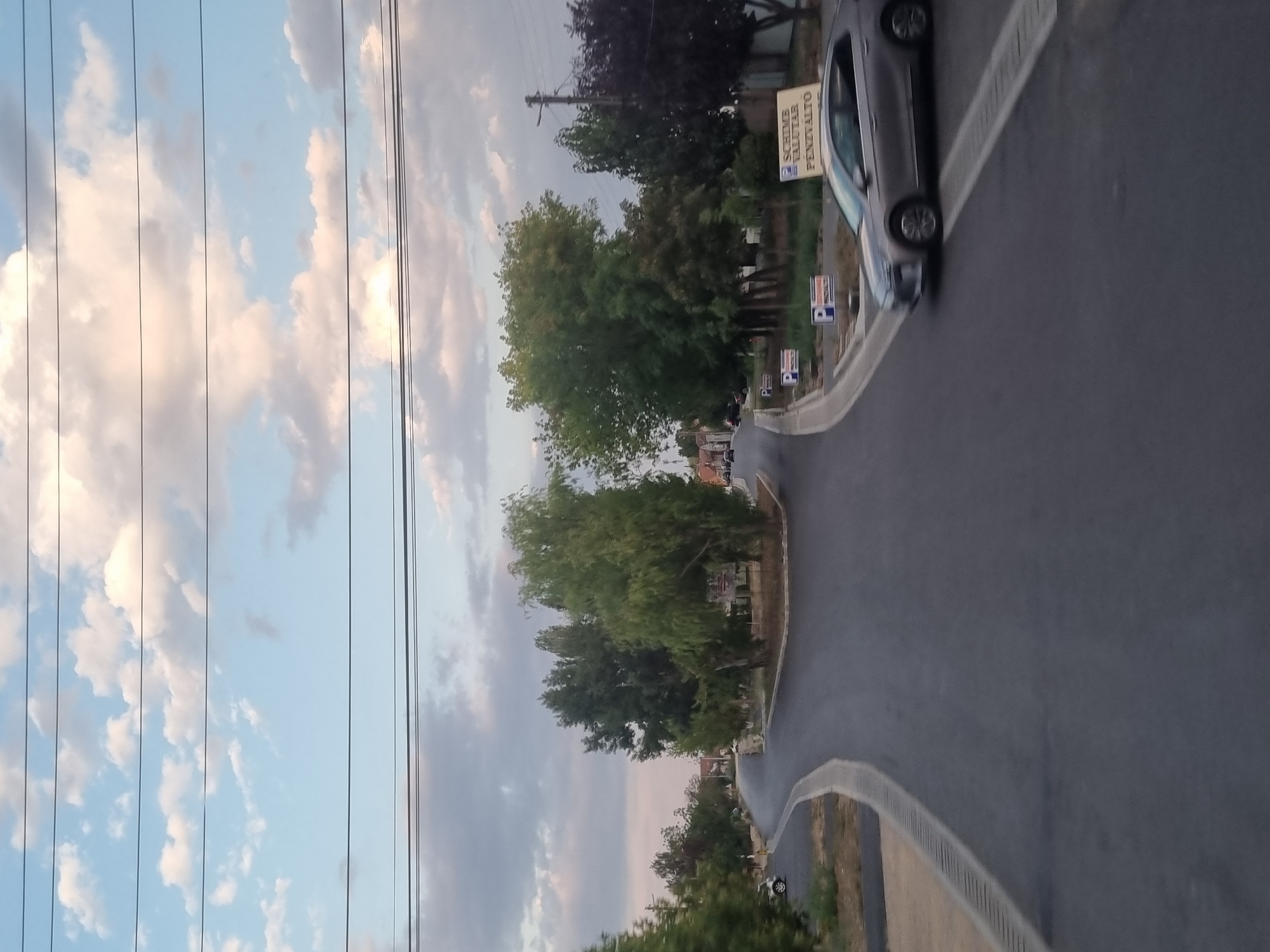 The image depicts a street intersection in Salonta, Romania, with a gray sedan driving on the right side of the road. The sky is partly cloudy, with a mix of blue sky and white and gray clouds. Power lines are visible crisscrossing the upper portion of the frame. Trees line the sides of the road, with some houses and buildings visible in the background. There are several parking signs, denoted by a "P" symbol, placed along the side of the road, and a larger sign that reads "SCHIME VALUTAR PENZVALTO". The road surface appears to be asphalt, and there are concrete curbs and drainage grates along the edges. The scene appears to be a typical suburban or semi-urban environment.