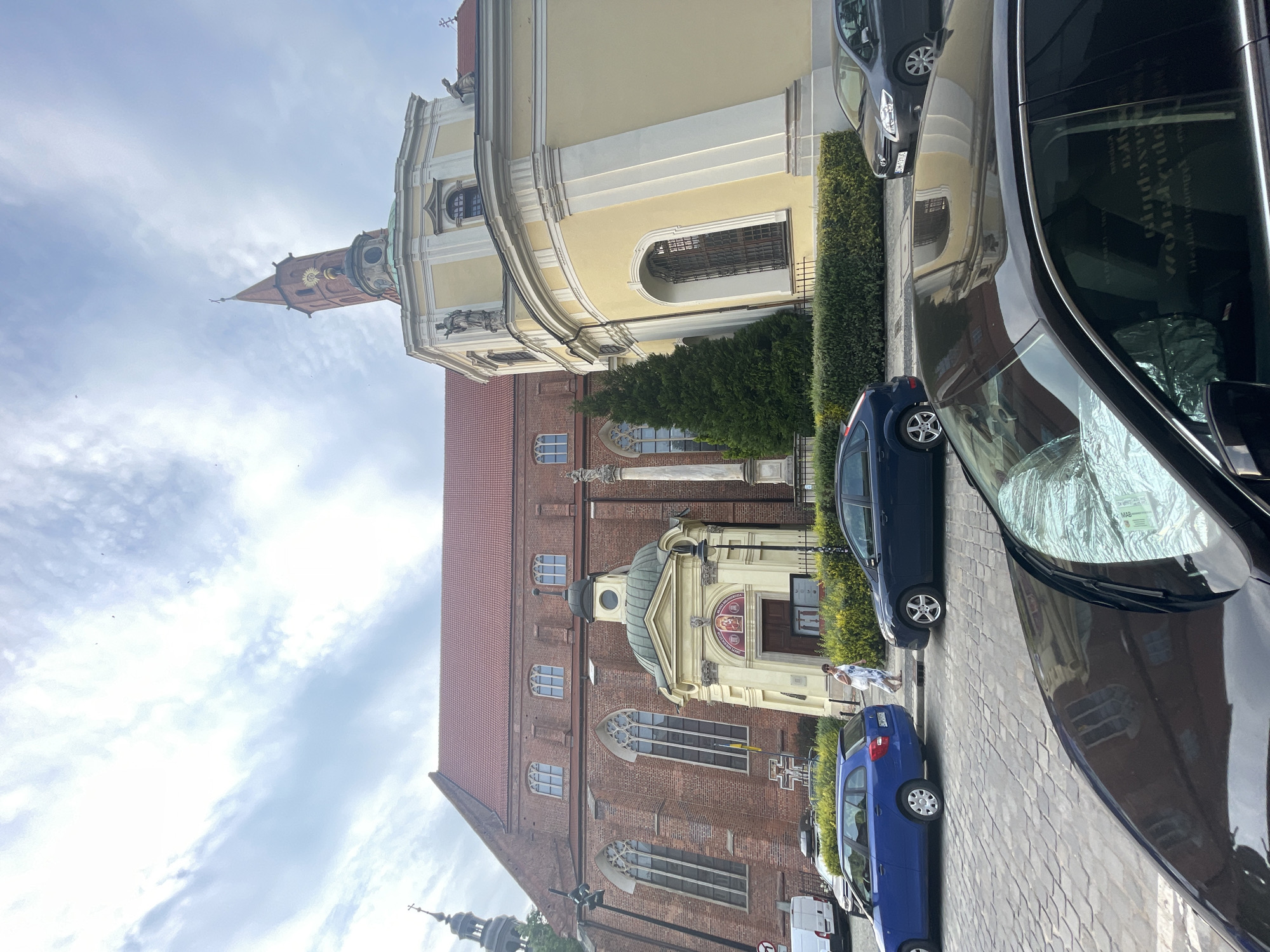 In Wrocław, Poland, a low-angle, eye-level view captures the exterior of the Cathedral of St. John the Baptist on a partly cloudy day. The main structure of the cathedral is a large, red brick building with a steeply pitched red tile roof and tall, arched windows. To the right of the main building, a lighter yellow, curved annex juts out, featuring a tall, red-domed tower topped with a spire and a cross. The annex is adorned with white trim and architectural details, and statues are visible on its upper level. In the foreground, a dark-colored car is parked, its windshield covered with a reflective sunshade. To the left, a blue station wagon and a dark blue sedan are parked on a cobblestone street. A woman in a light-colored dress walks away from the camera on the sidewalk. Dense green hedges line the area in front of the cathedral and annex.