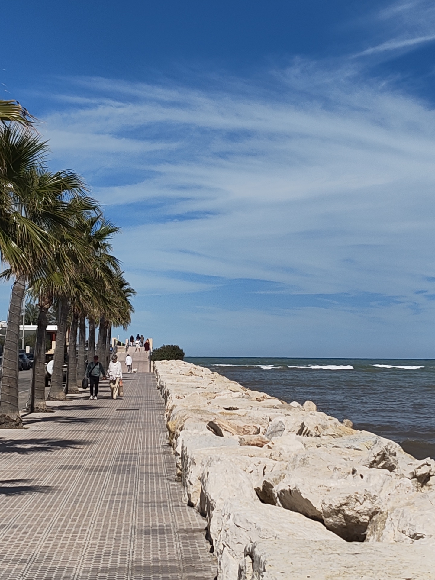 The image depicts a coastal scene in Dénia, Spain. A tiled walkway runs alongside a row of palm trees on the left and a stone barrier/breakwater on the right, separating the walkway from the sea. Several people are walking along the walkway, including a couple in the foreground and other figures further away. The sea is visible with small waves breaking, and the sky is partly cloudy with a mix of blue and white. Buildings and parked cars can be seen behind the palm trees on the left side of the walkway.