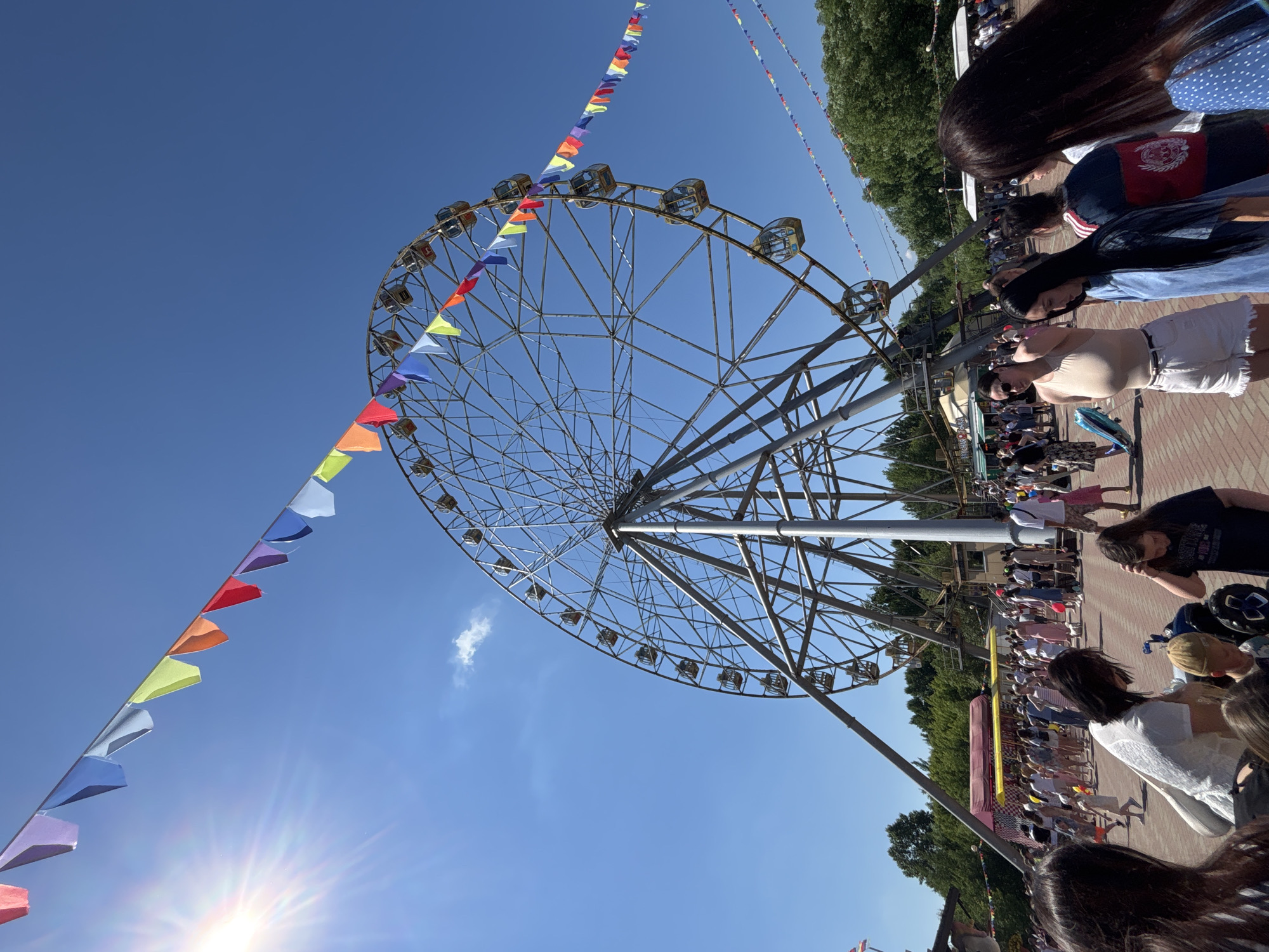 The image depicts a sunny day at an amusement park, likely in Rusanovo, Russia, with a large Ferris wheel dominating the background. Colorful triangular flags are strung across the bright blue sky, with the sun casting a strong glare on the left side of the frame. A crowd of people, including adults and children, are gathered on a brick pathway, milling around and engaging in various activities. In the mid-ground, there are food stalls or booths with awnings. Several Ferris wheel cabins are visible, and the structure of the Ferris wheel itself is intricate. The overall atmosphere appears to be festive and lively.