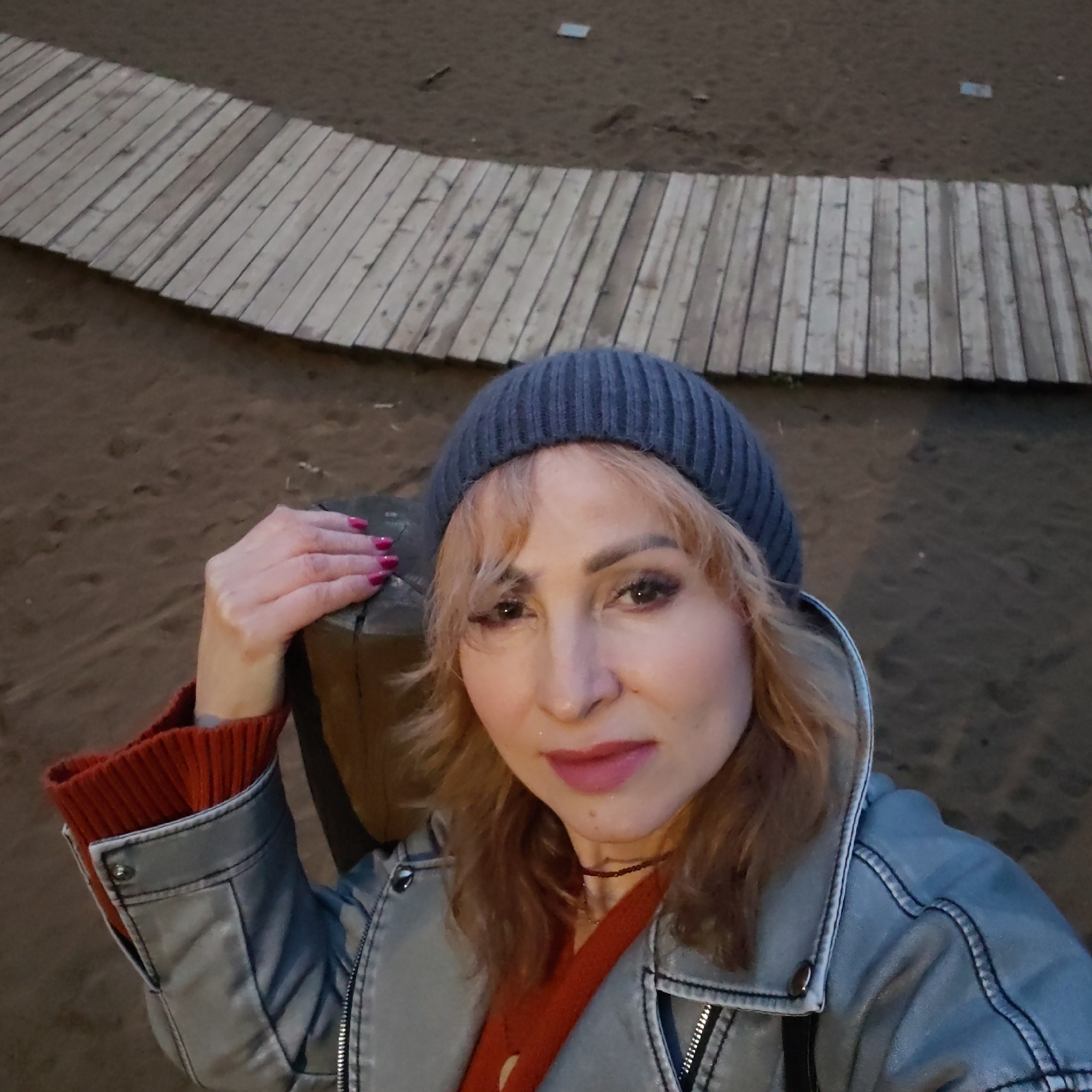 A woman stands on a sandy surface, likely a beach, with a wooden boardwalk behind her. She is wearing a gray beanie, a gray jacket, and an orange sweater with the sleeves visible. She is holding onto a wooden post. Her hair is blonde, and she has light makeup. Her fingernails are painted pink. Based on geolocation clues, it is probable that she is located in the city of Chaikovsky, Russia. The lighting suggests it may be evening or dusk.
